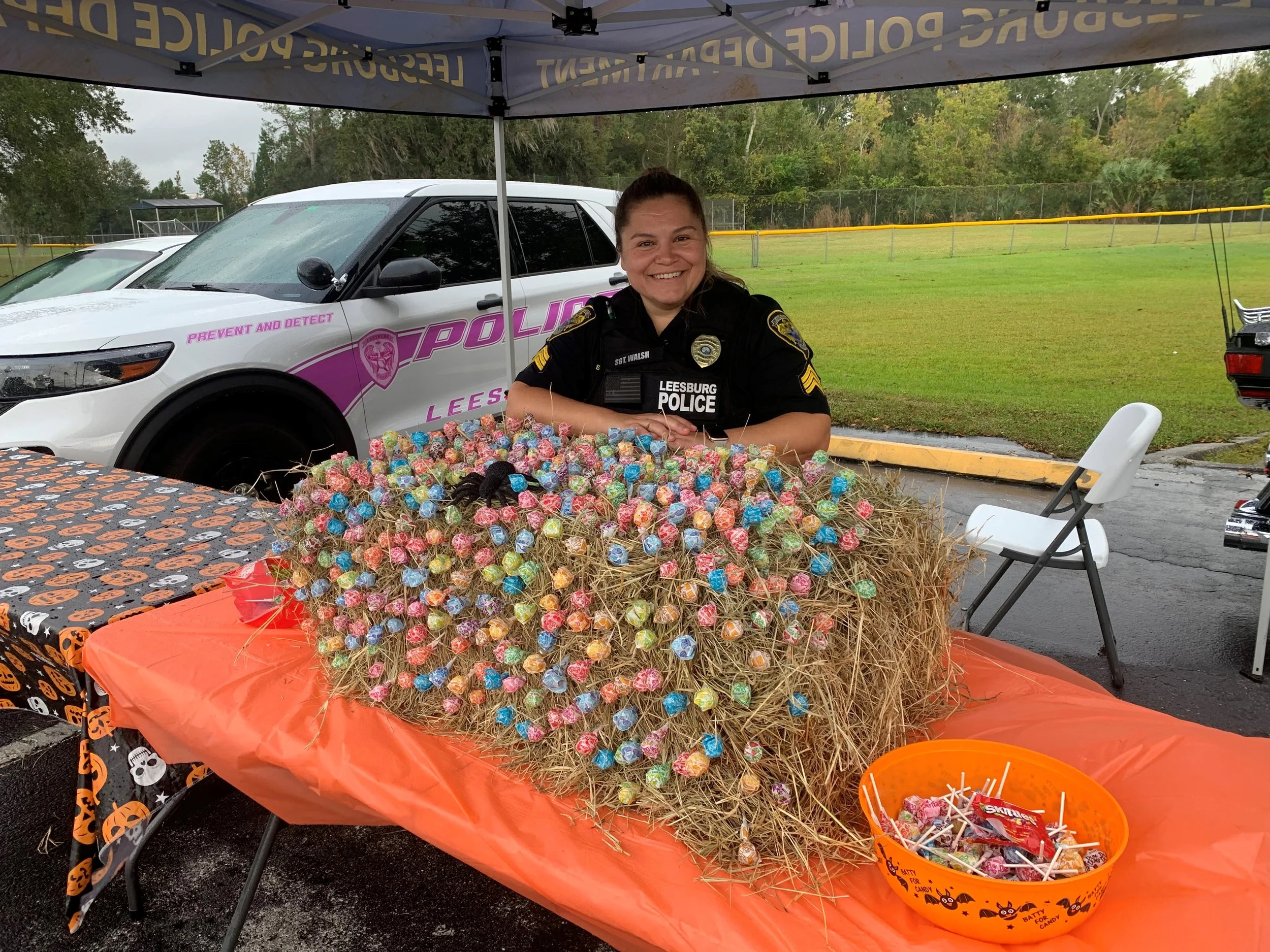 A police officer smiling behind a display of colorful lollipops on a table decorated with Halloween-themed tablecloths, outdoors under a tent with police vehicles in the background.