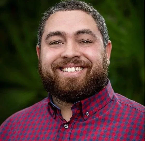 A man with a beard and short hair smiling outdoors, wearing a red and blue checkered shirt.