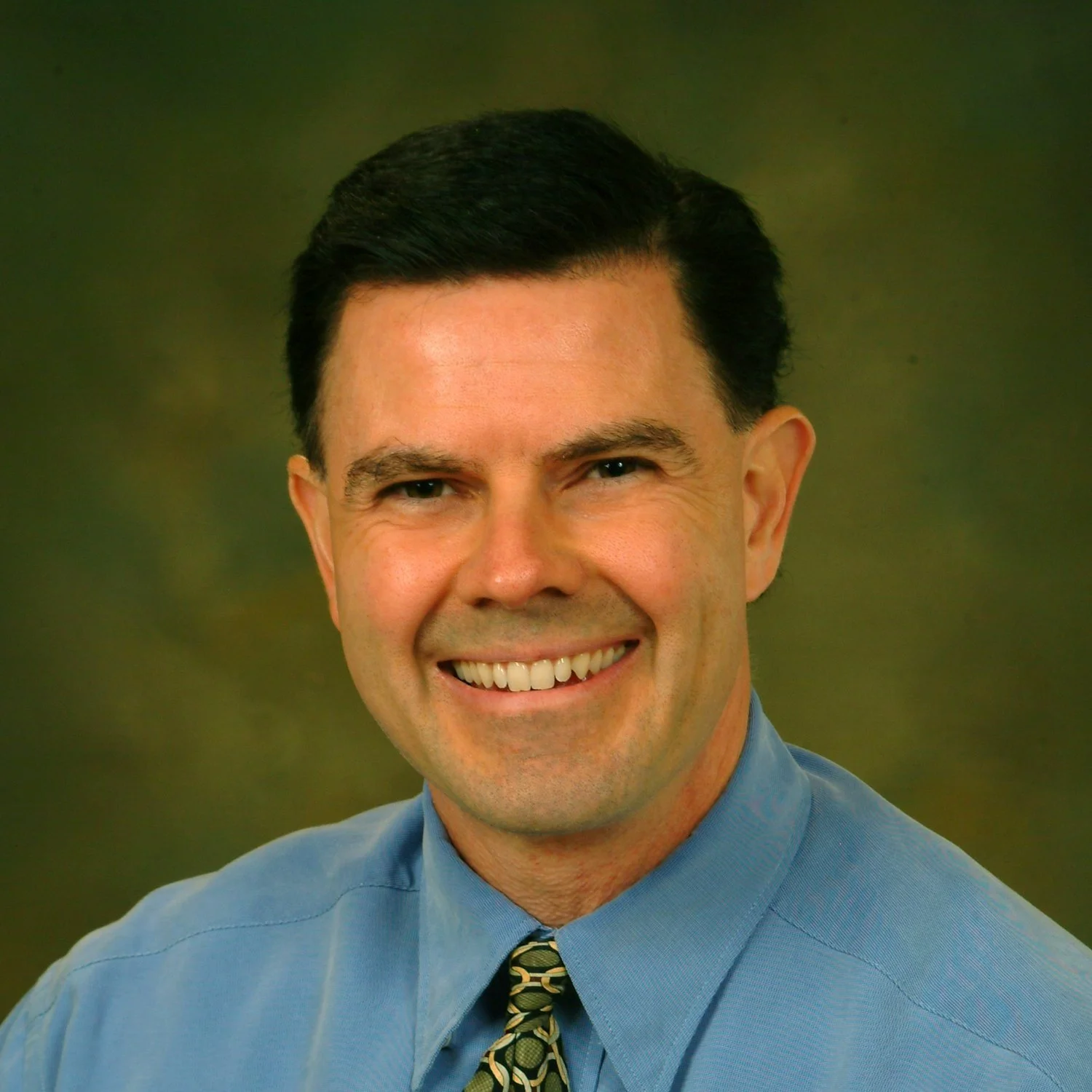 A smiling man with dark hair wearing a blue shirt and a patterned tie.
