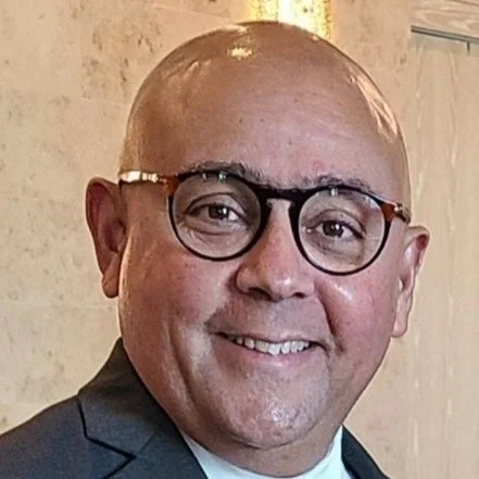 A smiling man with glasses and a suit, standing indoors.