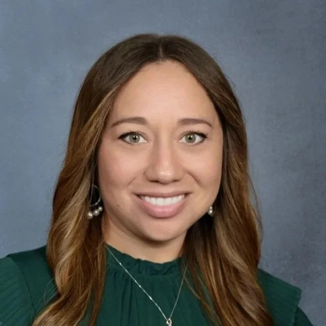 A young woman with long, wavy brown hair, wearing pearl earrings and a green top, smiling at the camera against a neutral background.