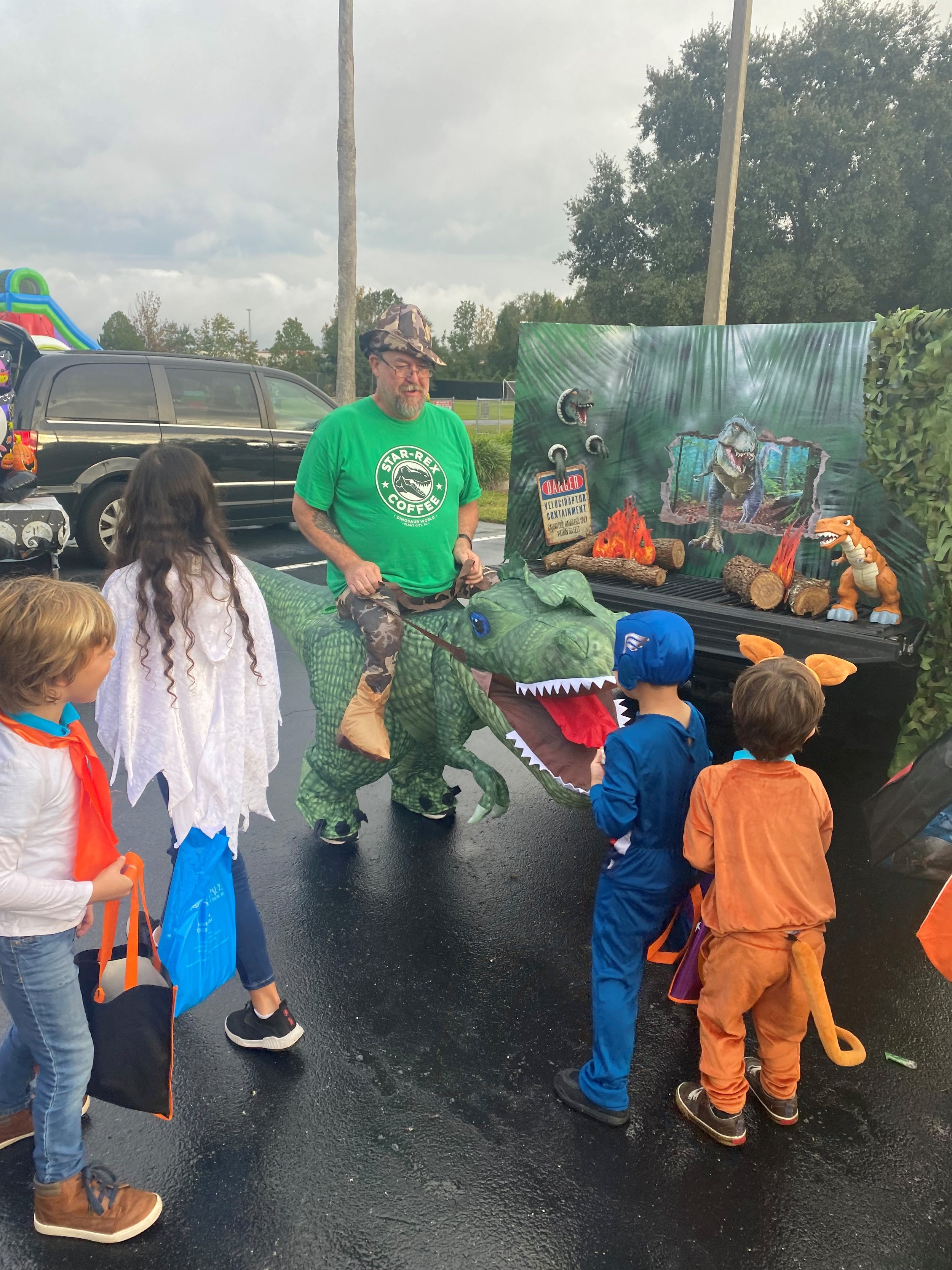 Man dressed as a dinosaur riding a green dinosaur costume, with children dressed in costumes, at a Halloween event in a parking lot with Halloween decorations and a small stage set with a forest scene, toy dinosaurs, and fake fire.
