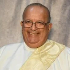 Smiling man wearing glasses and a white shirt with a cream-colored sash over his shoulder.