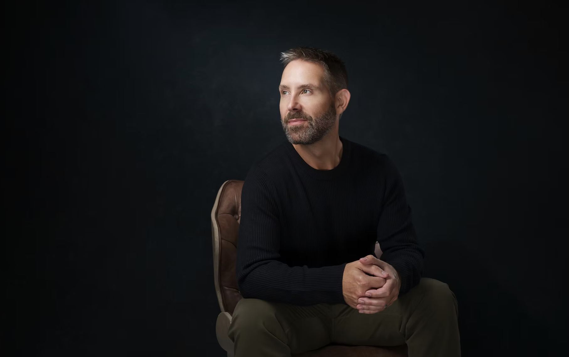 A middle-aged man with a beard and short hair, wearing a black sweater and khaki pants, sitting on a wooden chair with a dark background.
