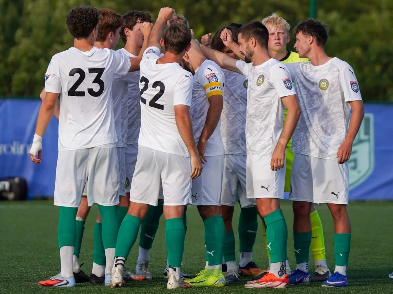 A group of soccer players in white jerseys and teal socks huddle together on the field, preparing for a game.