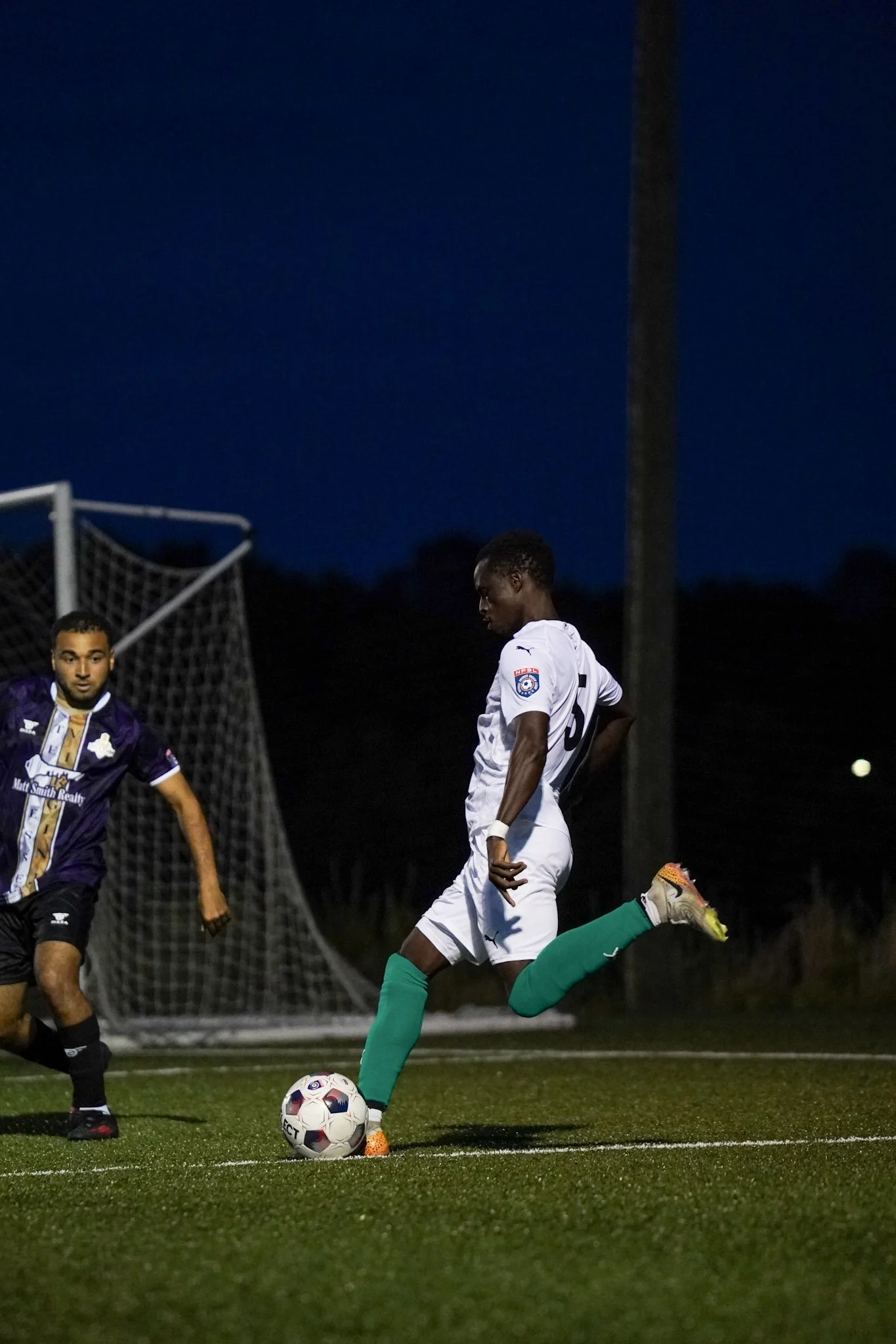 A soccer player in a white uniform with green socks kicking a soccer ball during a game at night, with another player in a purple jersey nearby and a goal net in the background.