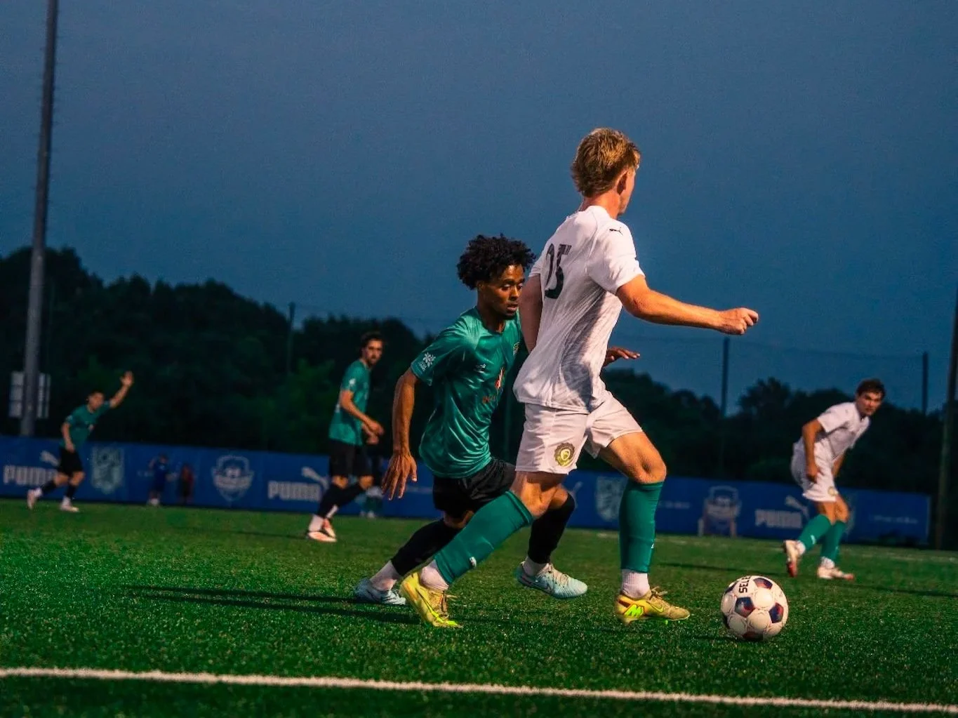 Soccer players competing for the ball on a field during a match at dusk.