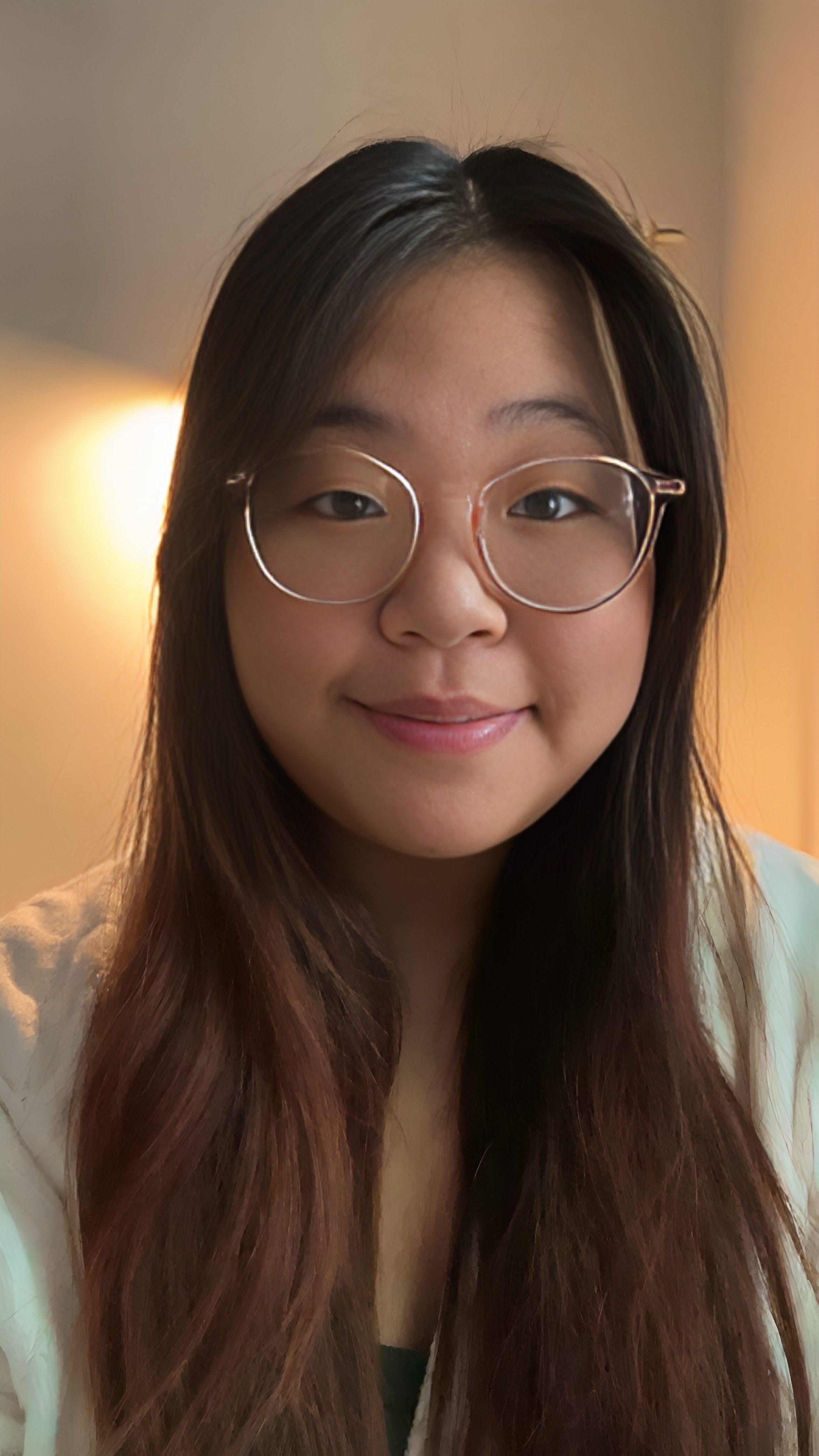 A young woman with long brown hair, glasses, and a gentle smile, standing indoors with a warm background light.