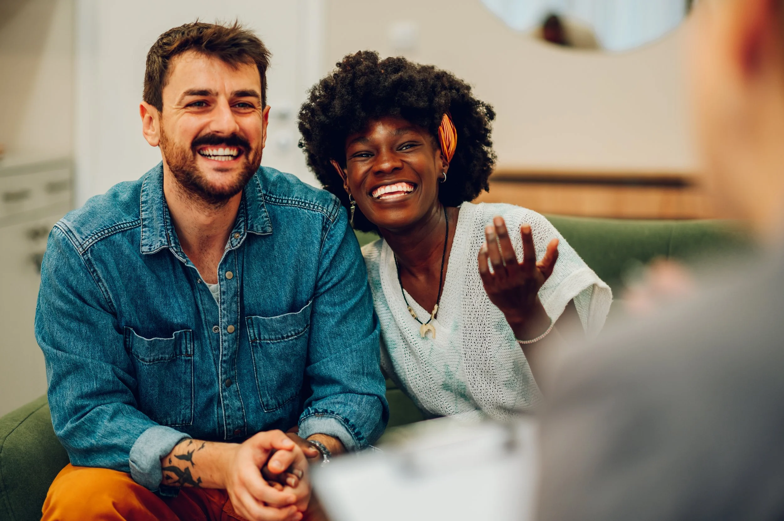 Two people, a man and a woman, sitting close together, smiling, and engaged in a conversation with someone not fully visible in the foreground.
