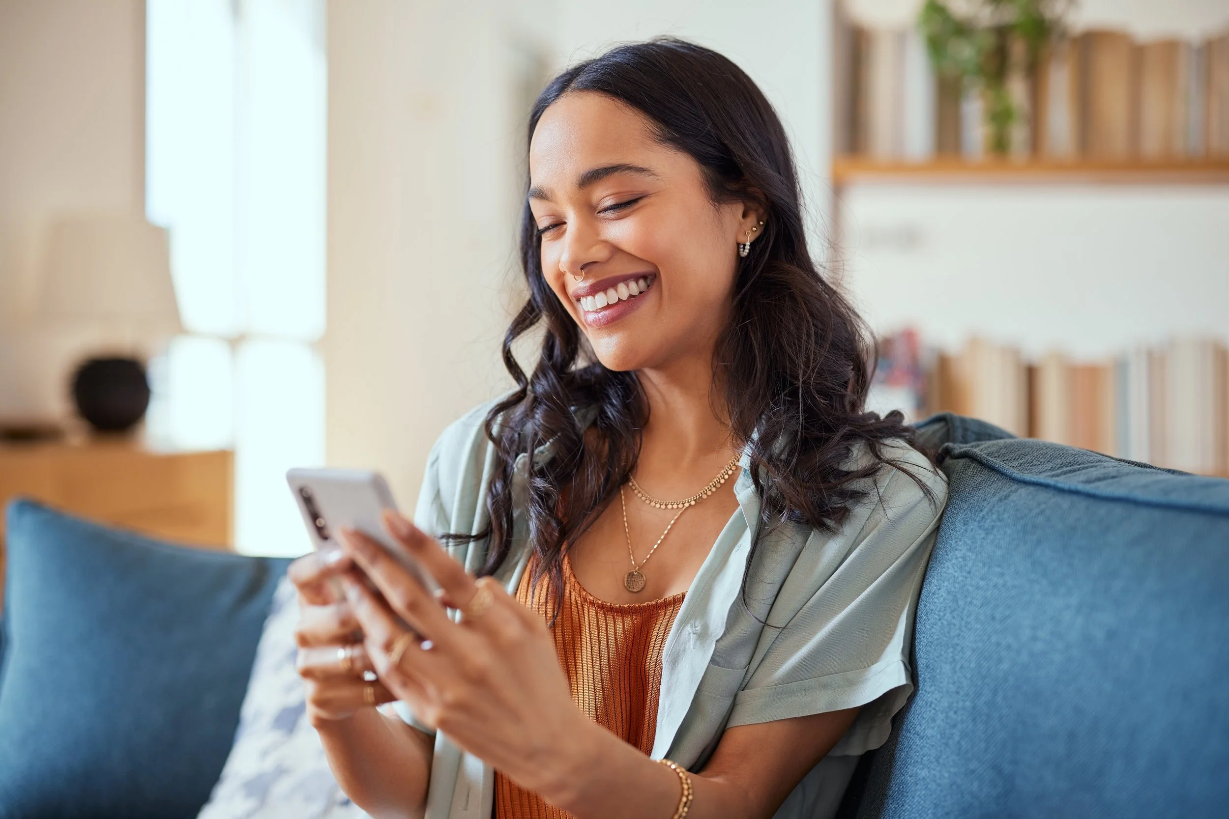 A young woman with dark wavy hair smiling while looking at her smartphone, sitting on a blue couch in a bright, cozy living room.