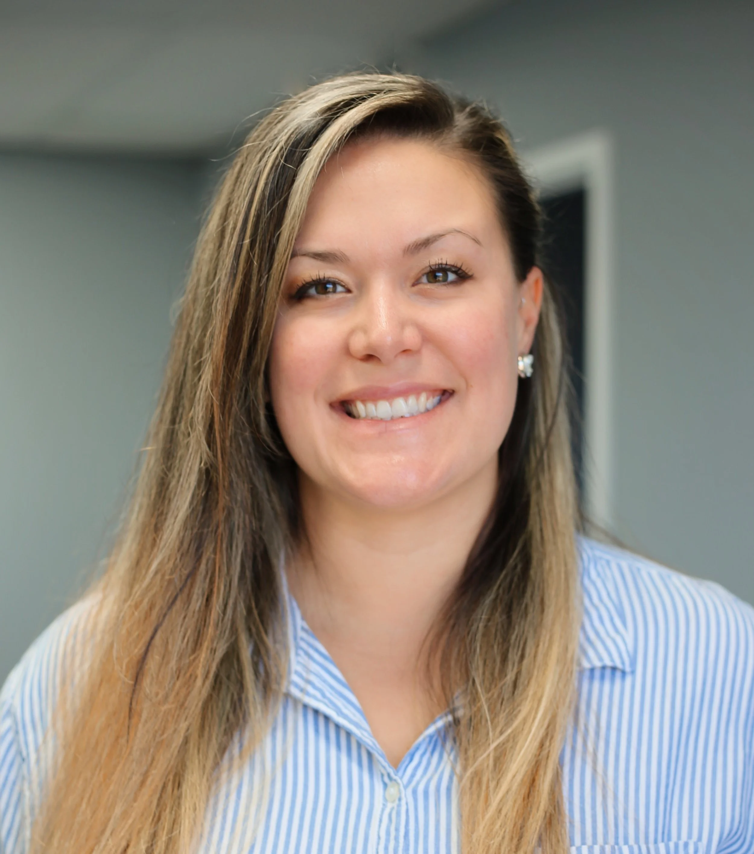 A woman with brown hair and light brown highlights, smiling, wearing earrings and a blue and white striped shirt.