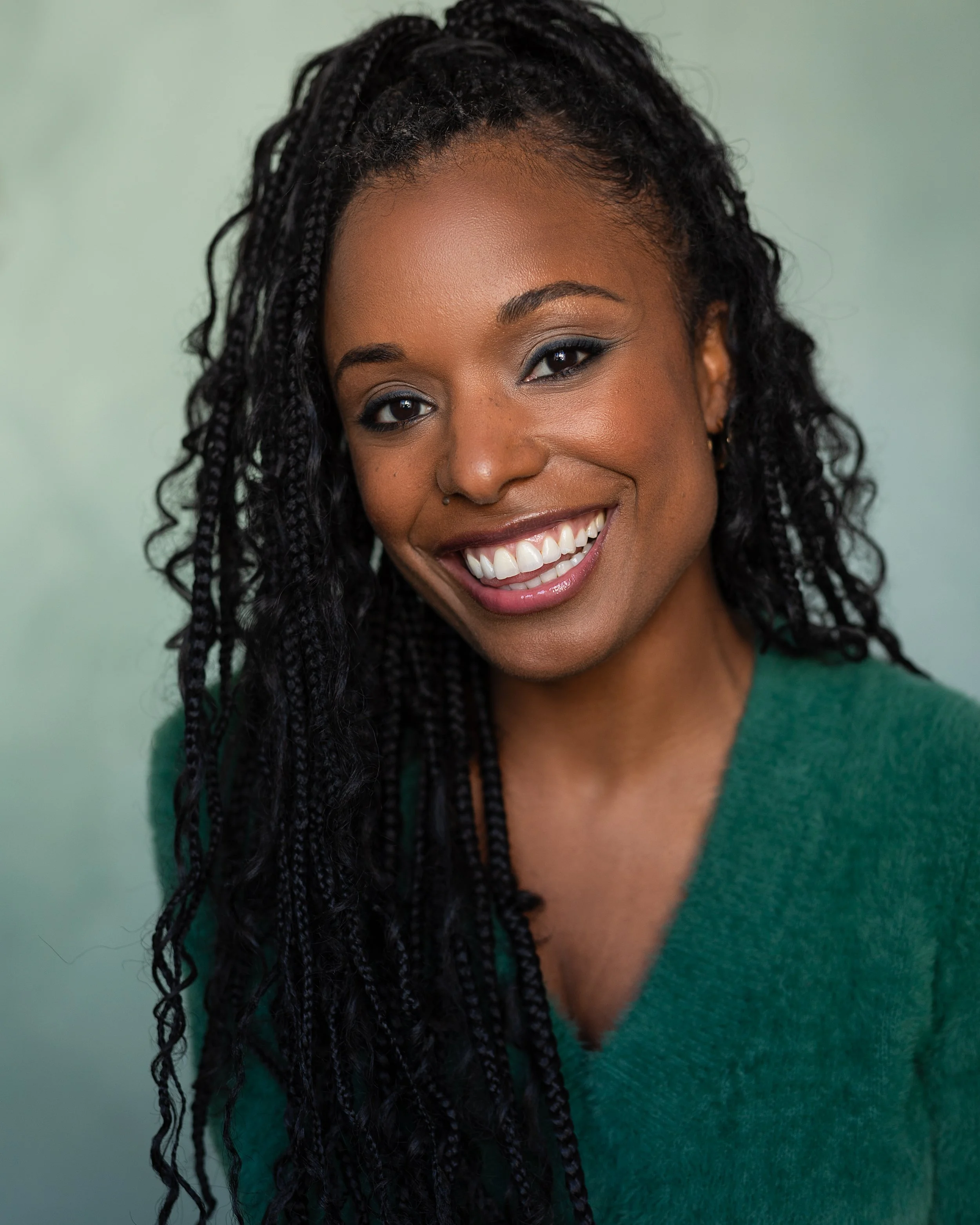 Close-up of a smiling African American woman with styled black braids and makeup, wearing a green top, against a light green background.