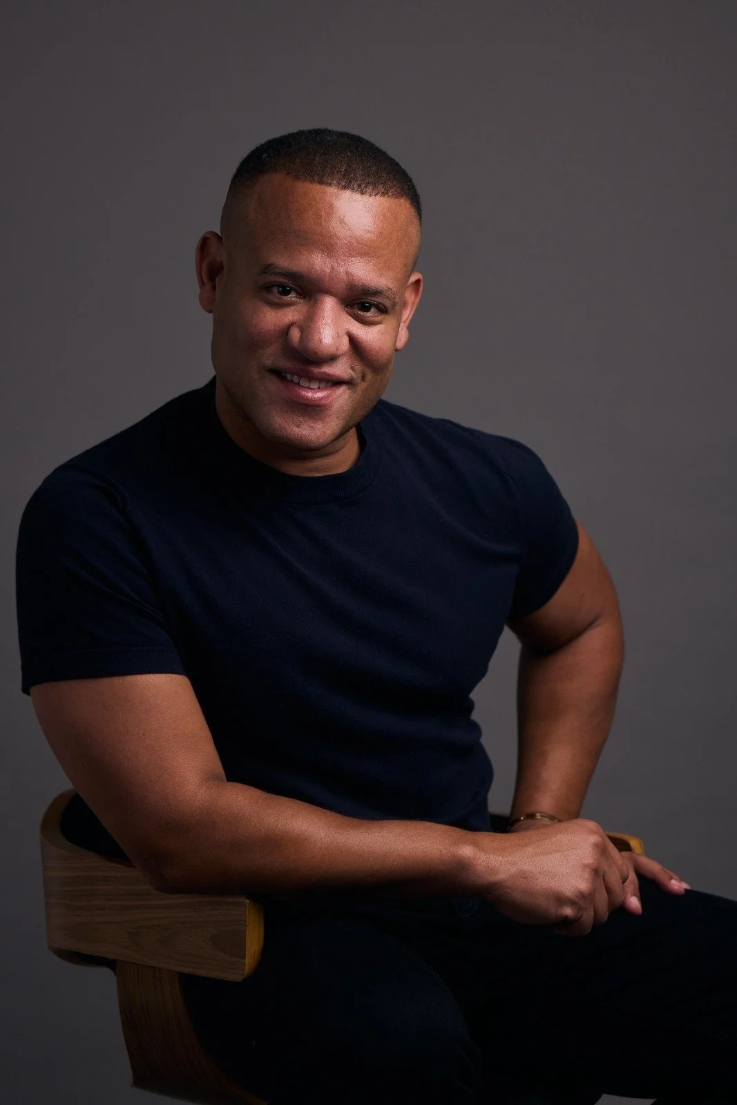 A man with short hair wearing a dark blue t-shirt, smiling and sitting on a wooden chair against a gray background.