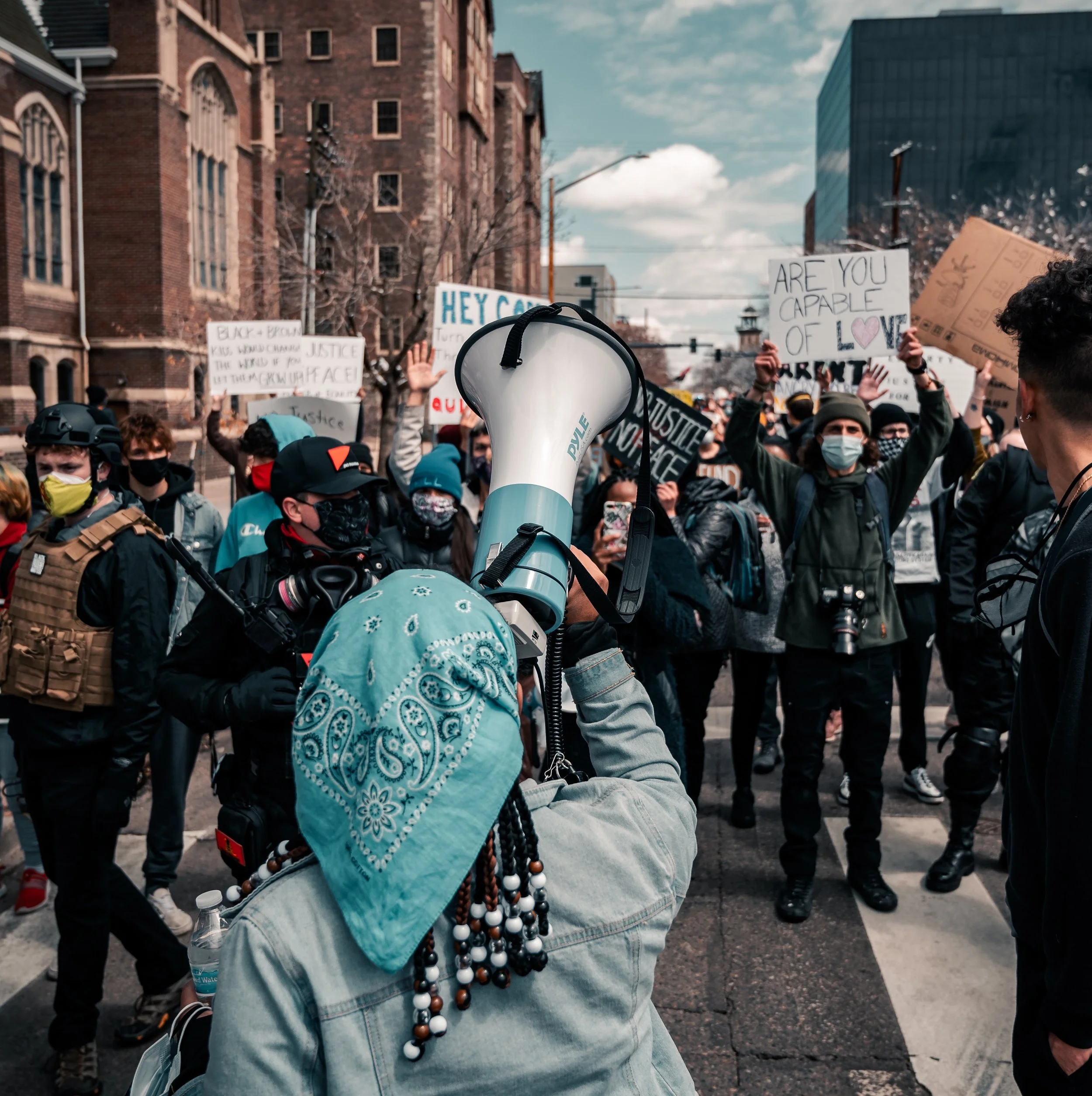 Crowd of people protesting on the street, many holding signs and banners with messages about justice and love, wearing masks and warm clothing.