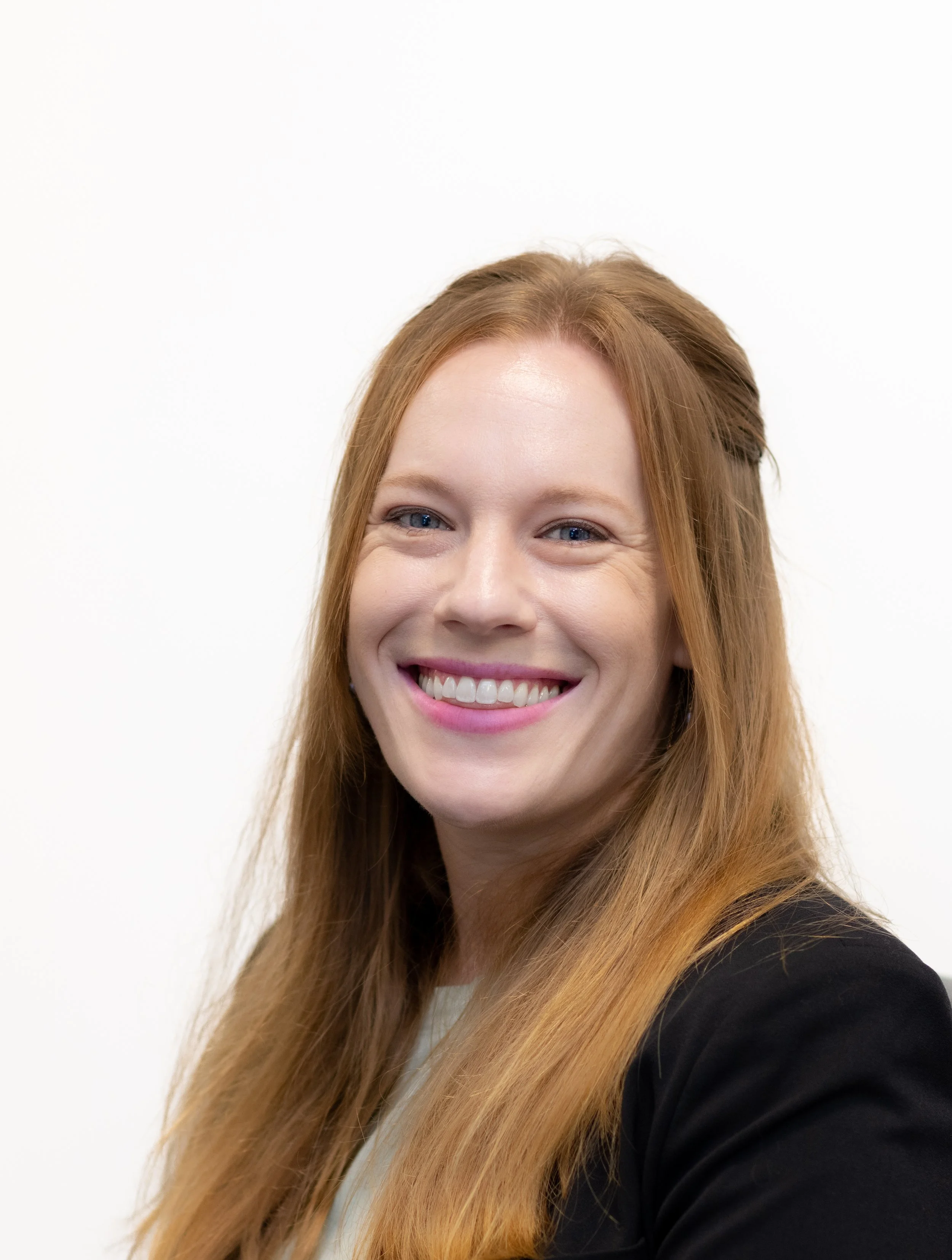 Image of woman with long red hair and bright smile with black blazer with a plain white background