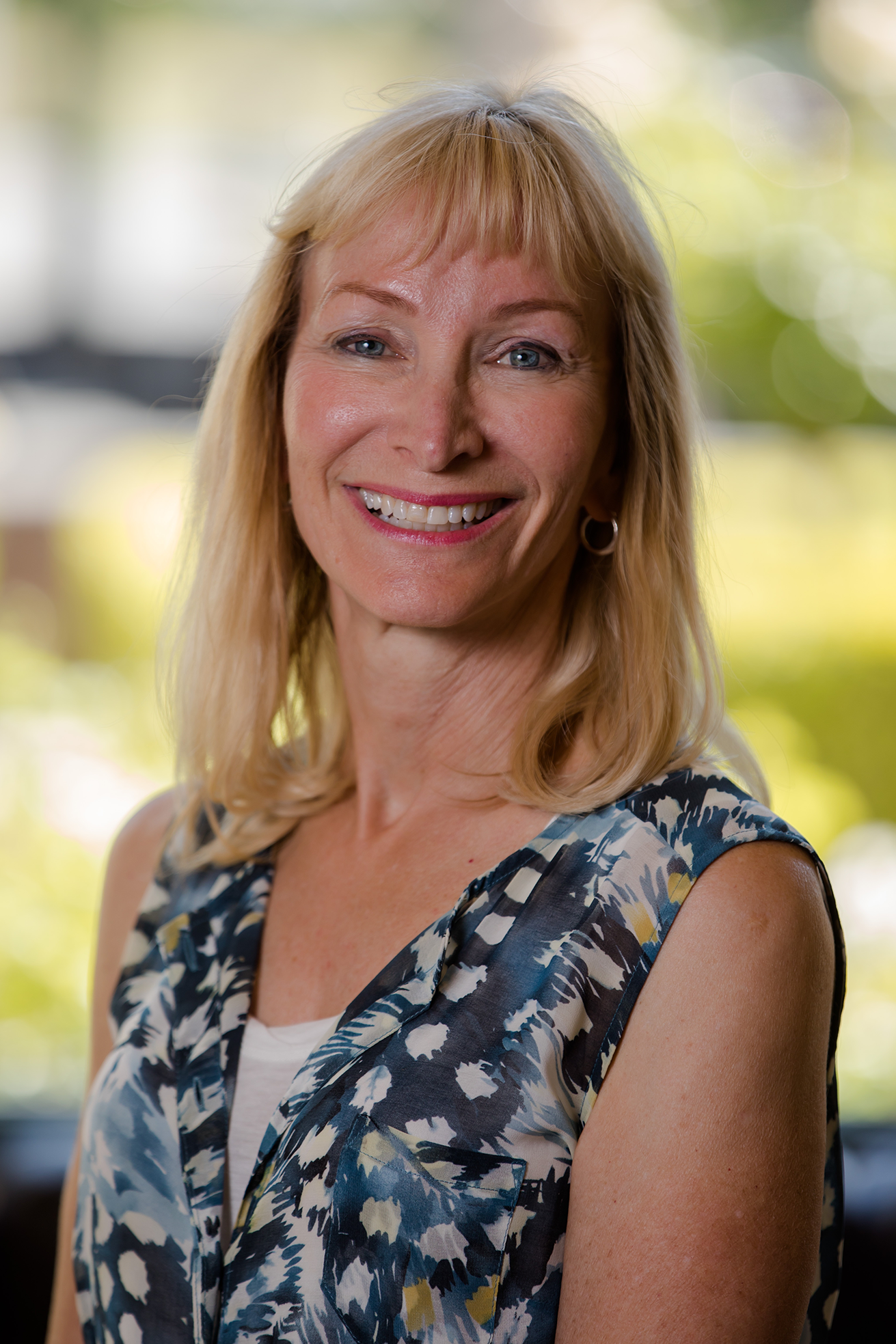 A woman with short blonde hair with bright smile in front of a blurred green nature background