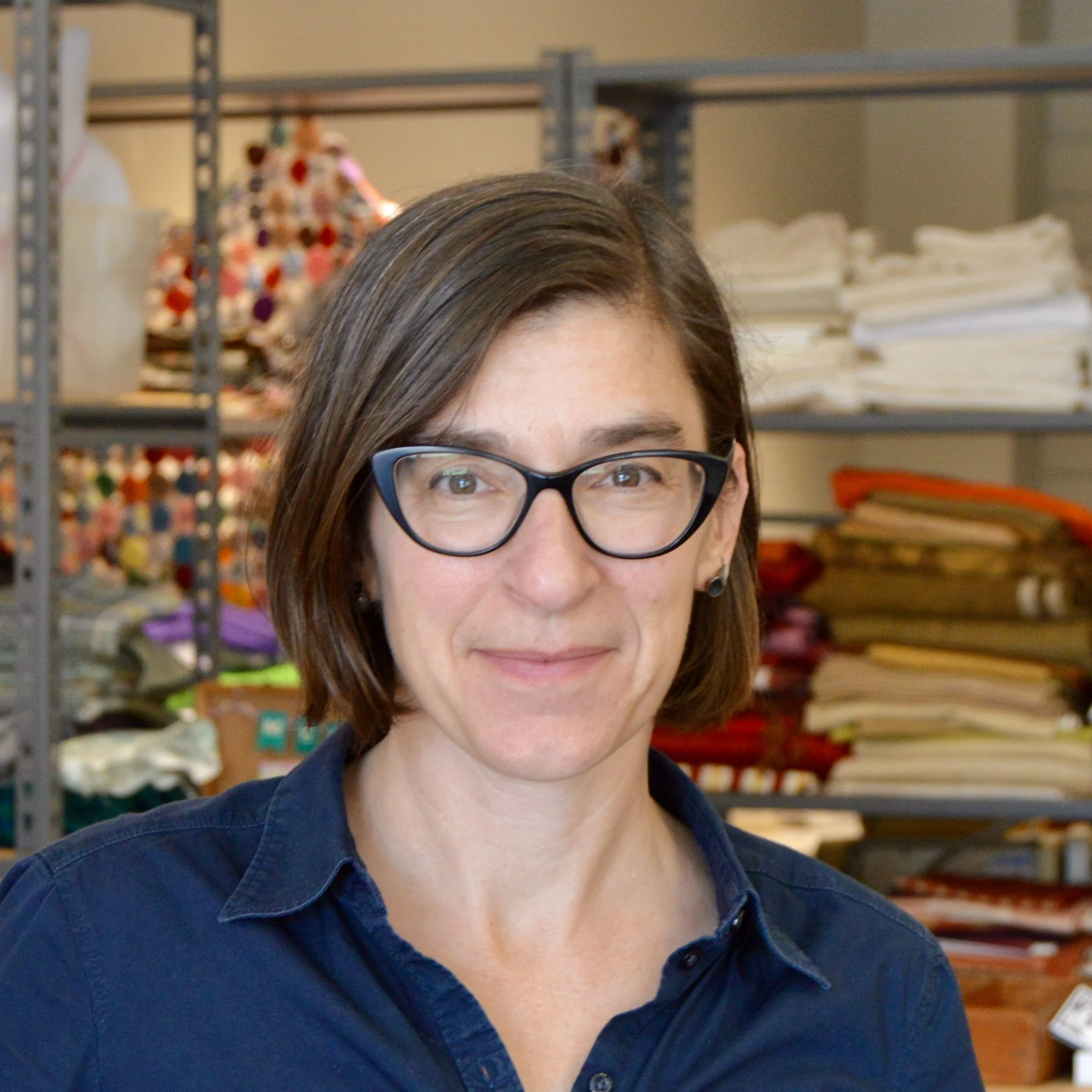 Image of a woman with short brown hair in a navy blue collared shirt in front of shelves with folded colorful fabric.