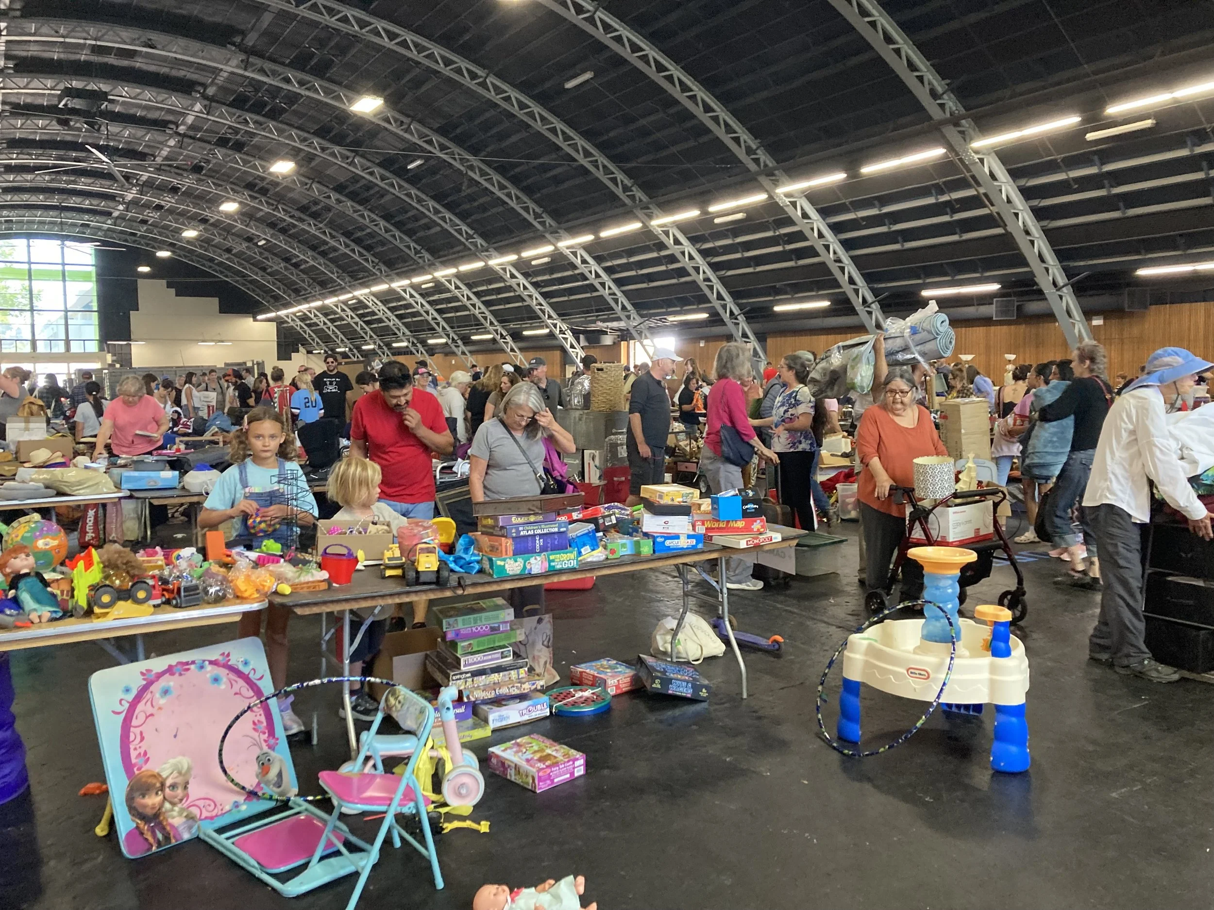 Large room with vaulted ceilings featuring tables with kids toys and dozens of people looking through items