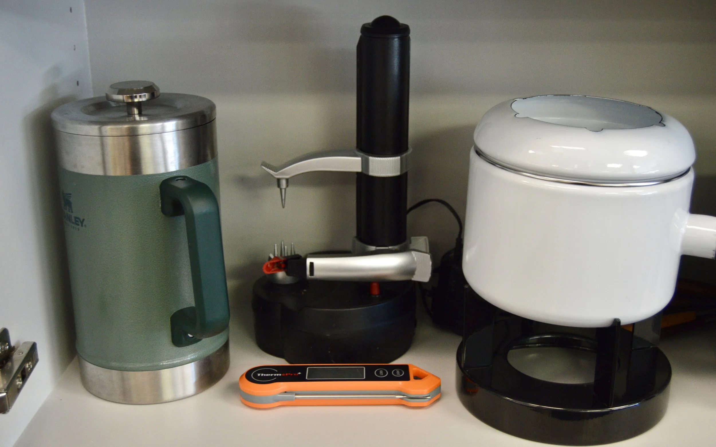 Cabinet shelf a silver and green thermos, a black electric peeler, and a digital thermometer in an orange case.