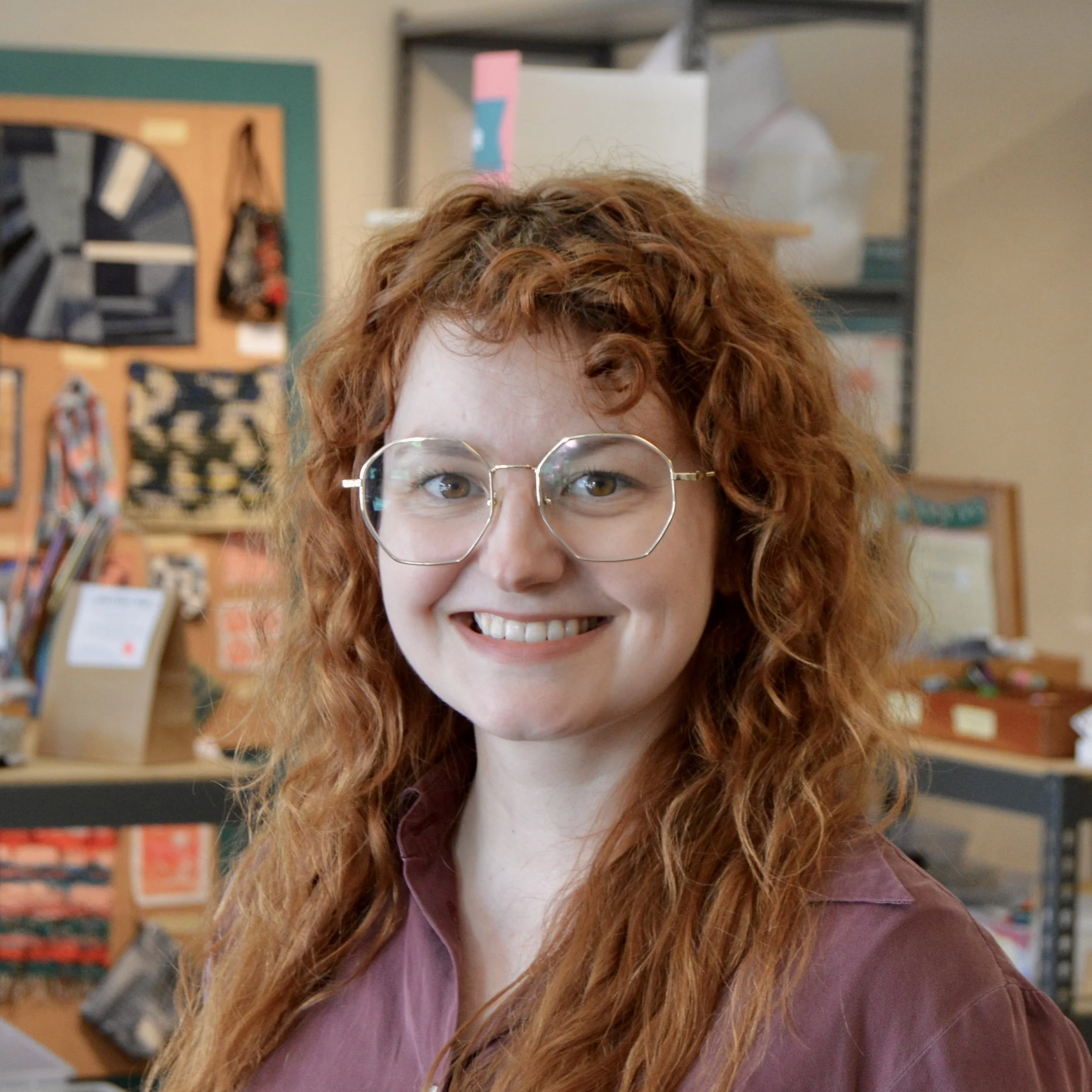 Image of woman with long red curly hair and silver glasses in a mauve collared shirt in front of shelves and wall hangings of textile projects and supplies