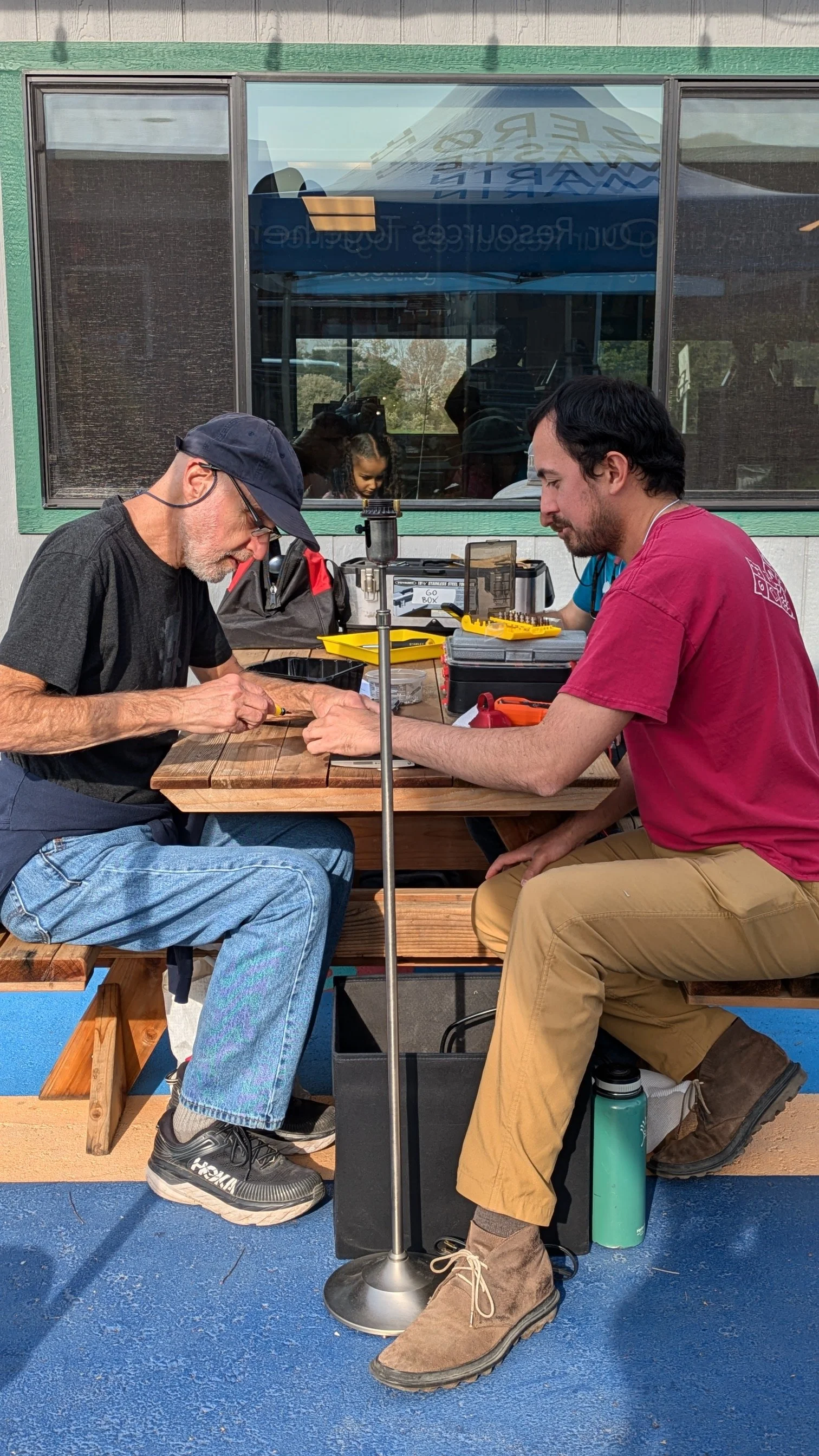 Two men working on electronics or small devices at an outdoor wooden table. One man is elderly with a gray beard, wearing a black shirt, jeans, a cap, and glasses. The other is younger with dark hair, wearing a red shirt, tan pants, and brown work boots. There are tools and electronic parts on the table, and a window behind them reflects other people and outdoors.
