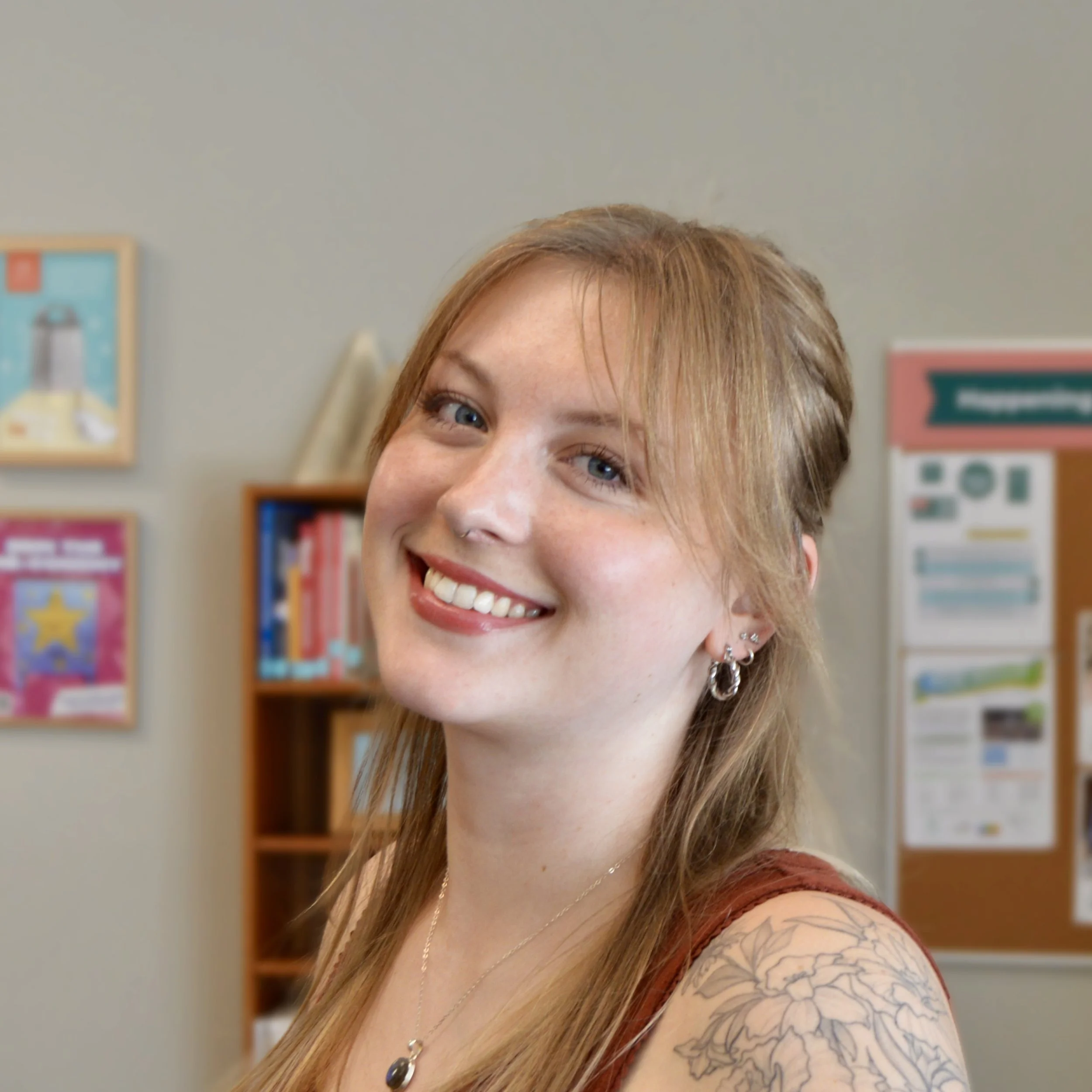 Image of woman with medium length blond hair with bright smile in front of grey wall with bright posters and bookshelf.