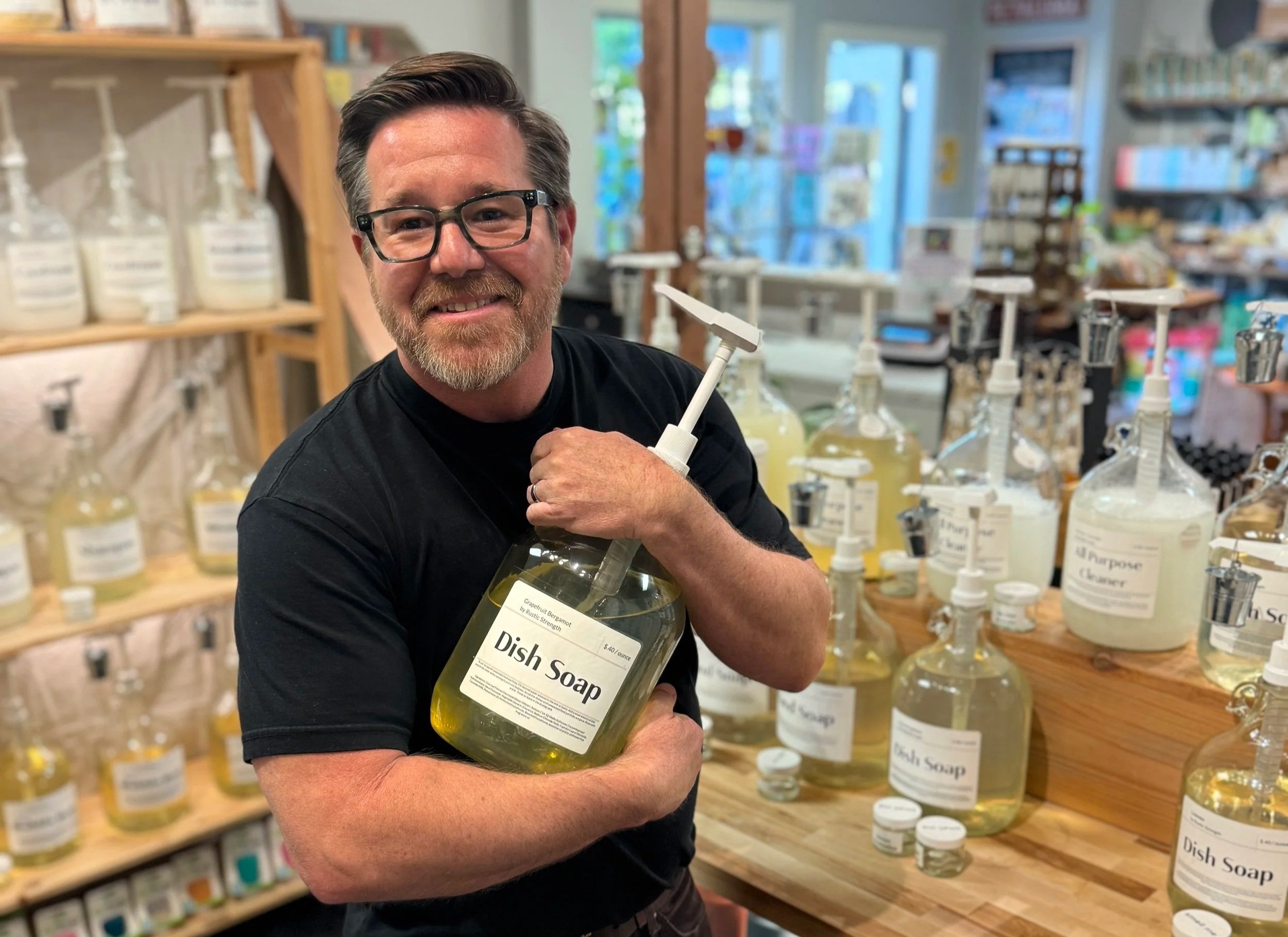 Man clutching to large bottle labeled "dish soap" with other various large bottles in background