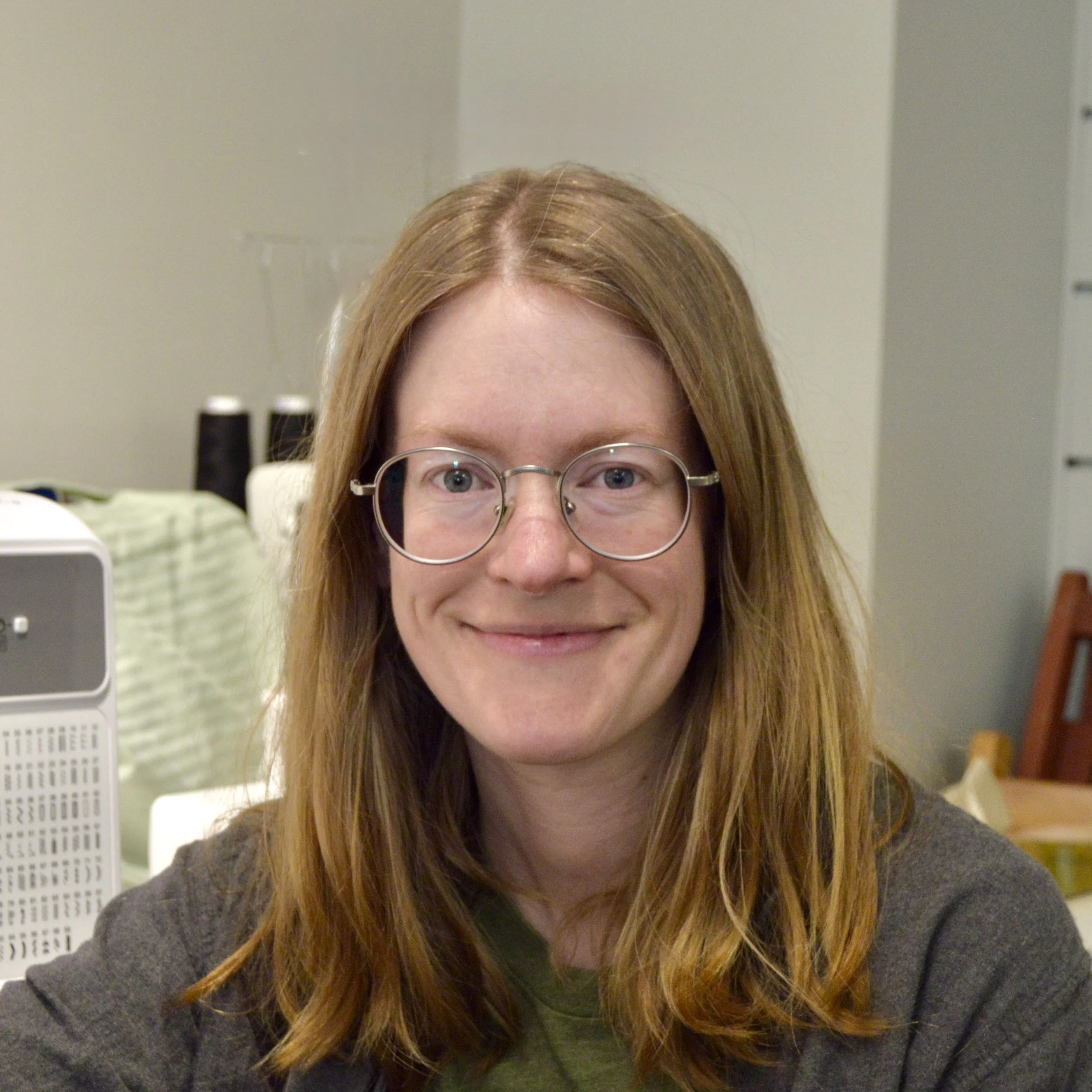 Woman with glasses with closed mouth smile in grey top with plain grey walls as background