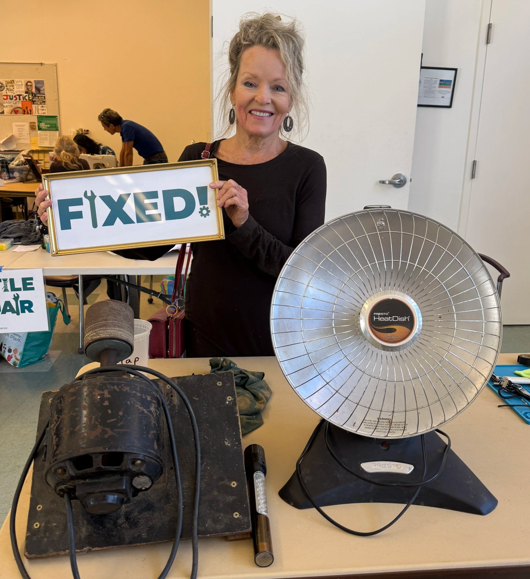 An older woman holding a framed sign that reads 'FIXED!'. She is smiling and standing behind a table with old fan parts, including a fan motor and a  heat dish. In the background, there are people working at a table and a colorful poster on the wall.