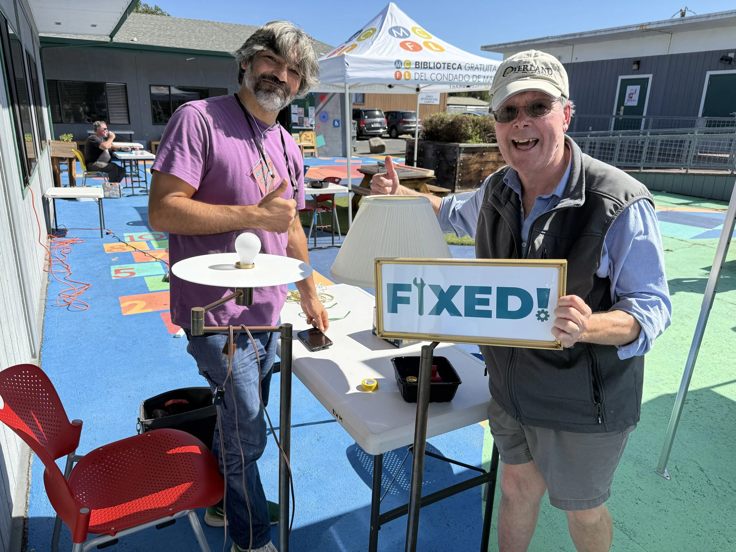 Two people with big smiles and hands in a thumbs up position. Person on the right is holding a "fixed" sign.