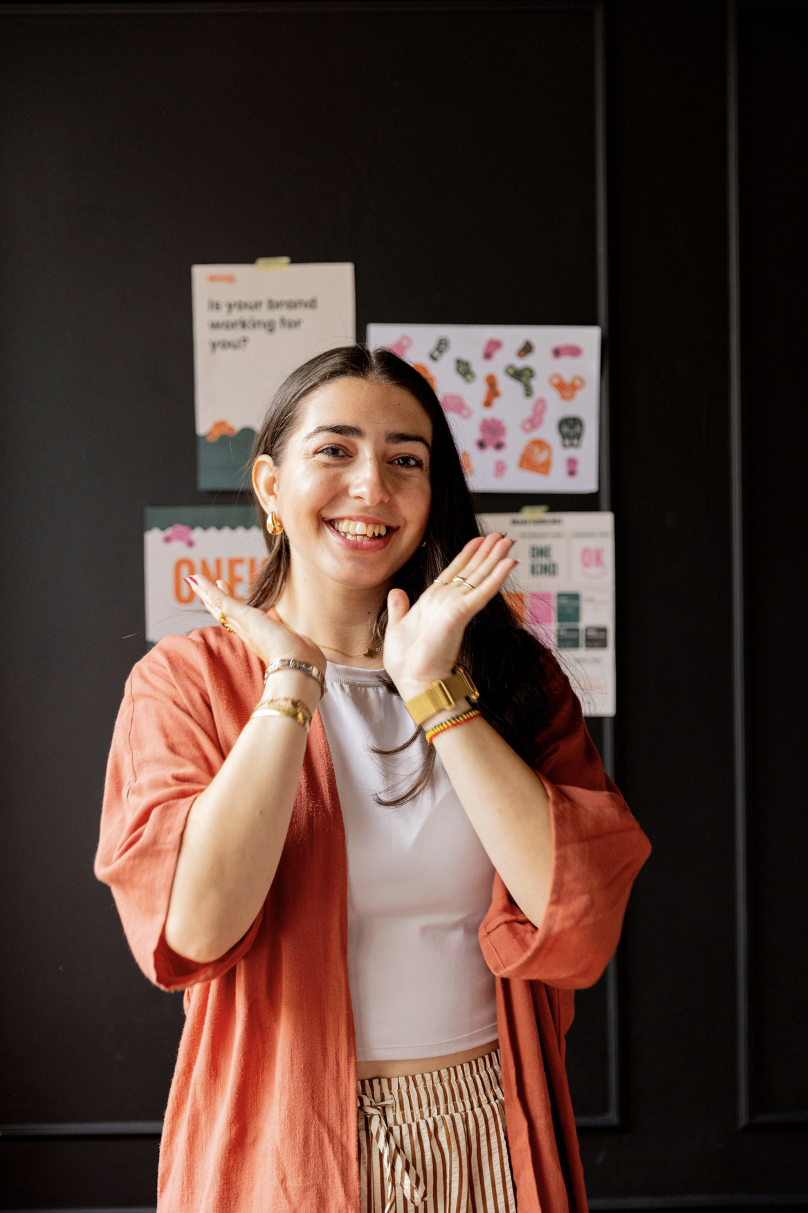 Woman with dark hair and bright smile with hands framing face wearing a white and orange top with a black background with colorful posters.