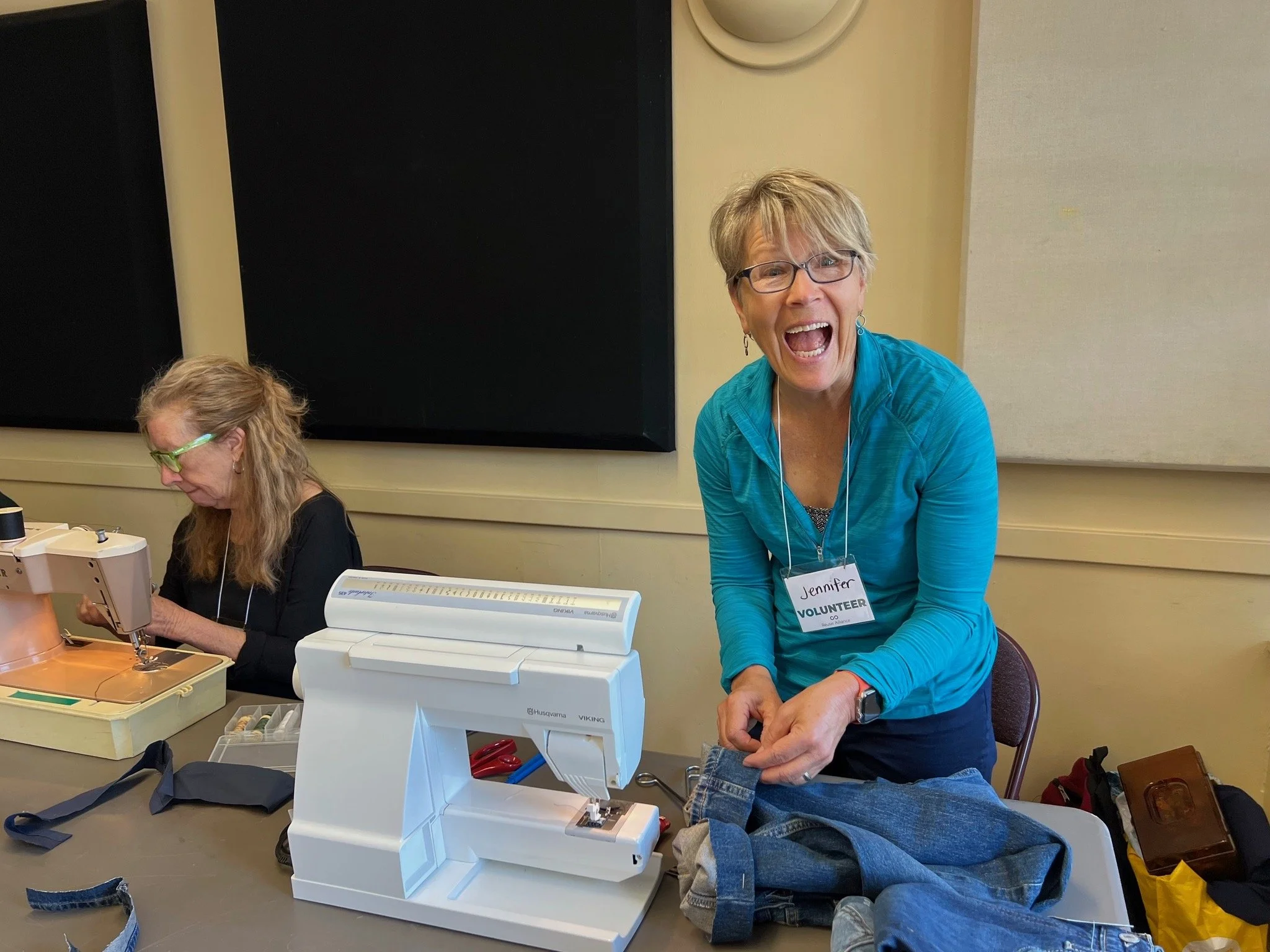 A woman with short blonde hair, glasses, and a blue jacket is smiling and wearing a volunteer badge, standing at a sewing or crafting event. Another woman with long blonde hair and glasses focuses on her project at a sewing machine on a table.