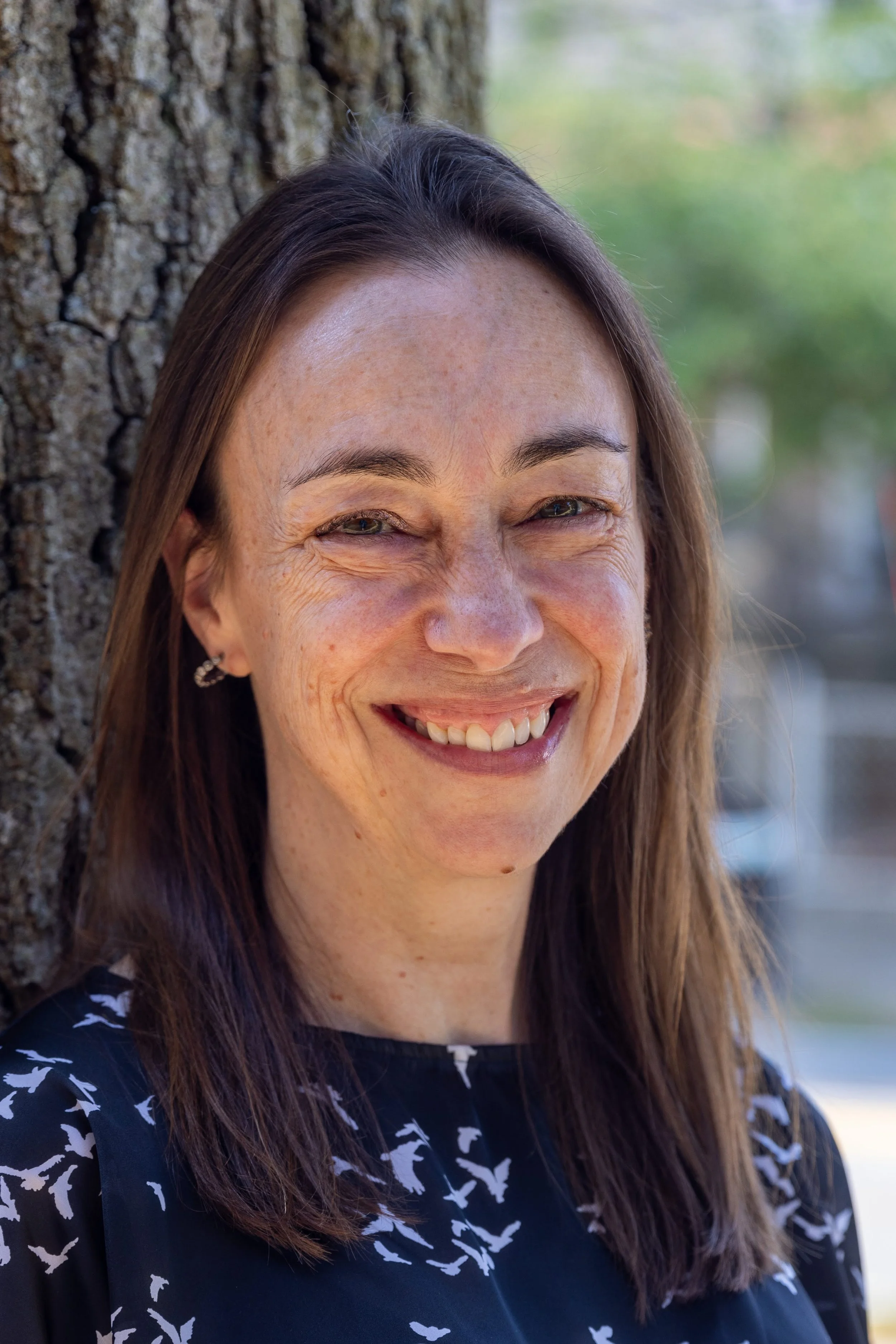 A woman with long brown hair, fair skin, and freckles, smiling and leaning against a tree in an outdoor setting with blurred greenery and sky in the background.