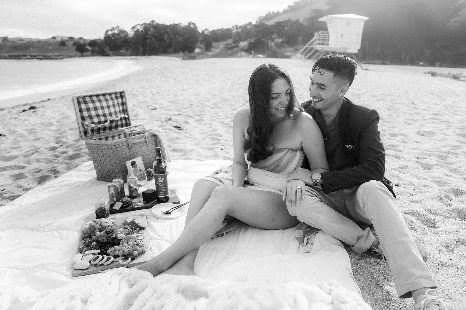 A couple enjoying a picnic on the beach, sitting on a blanket with a spread of food, drinks, and snacks, while smiling and looking at each other.