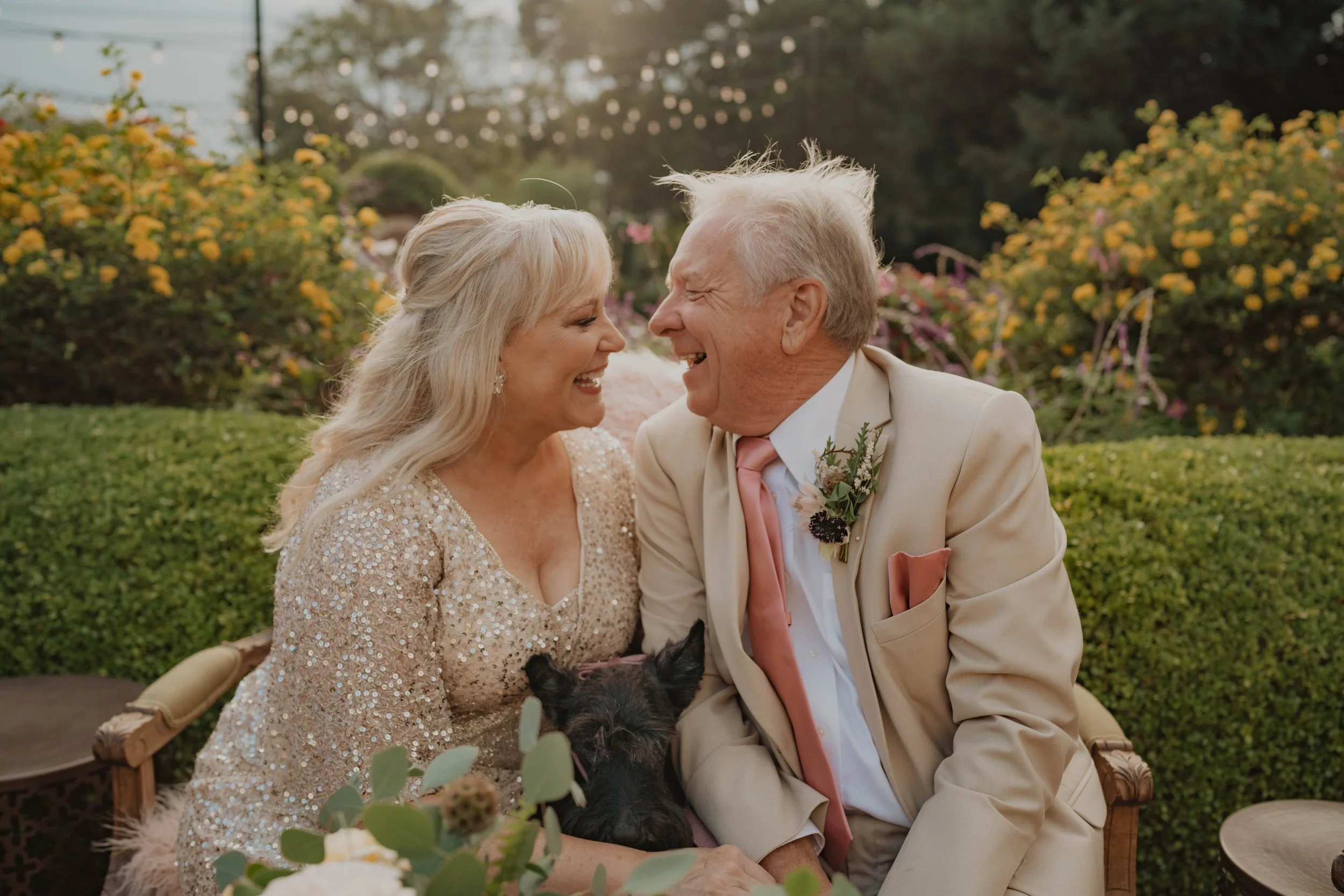 An elderly couple sitting outdoors in a garden, laughing and holding a small black dog, dressed in formal attire, with string lights and flowers in the background.