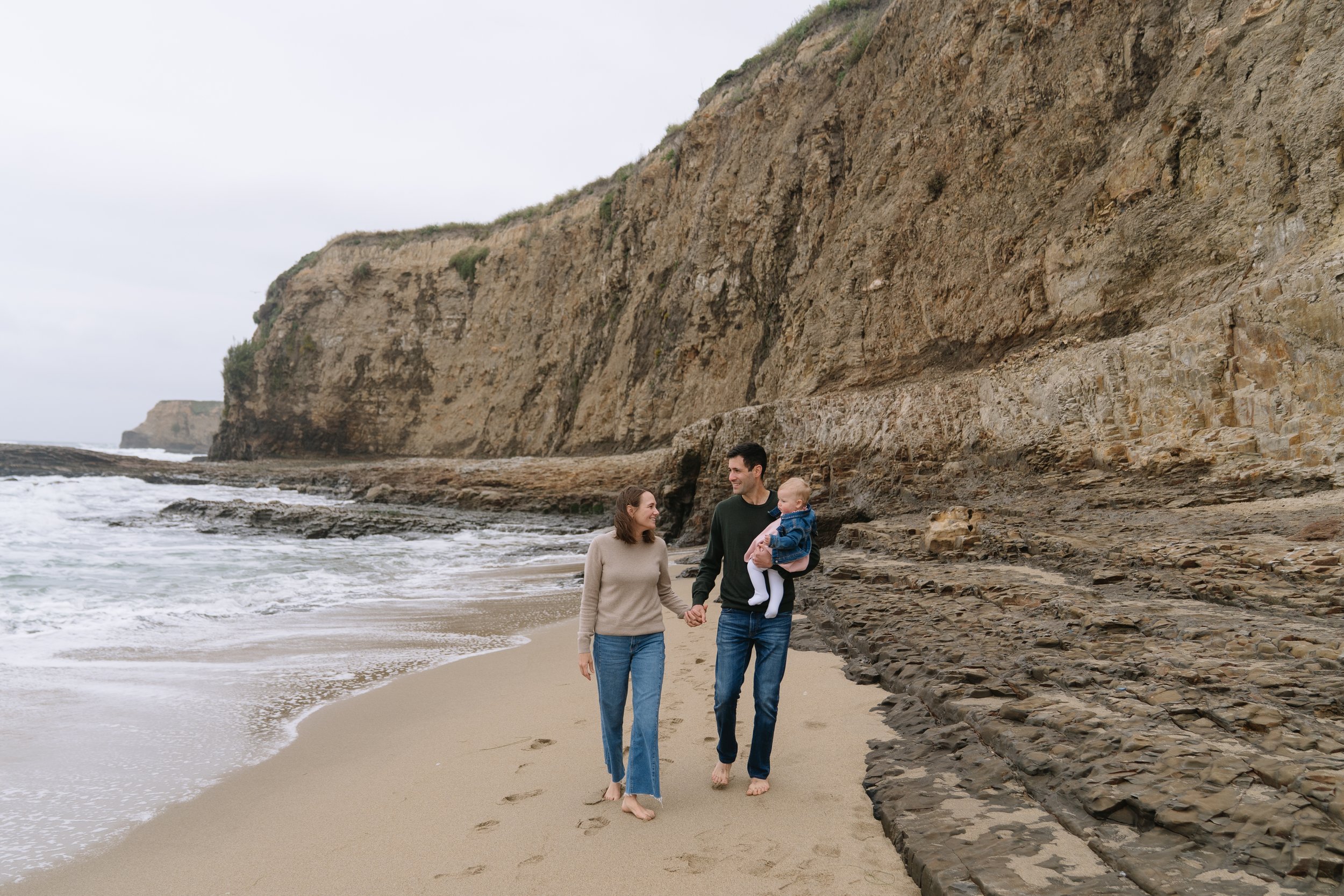 A family of three walking barefoot on a sandy beach near rocky cliffs, holding hands and smiling, overcast sky.