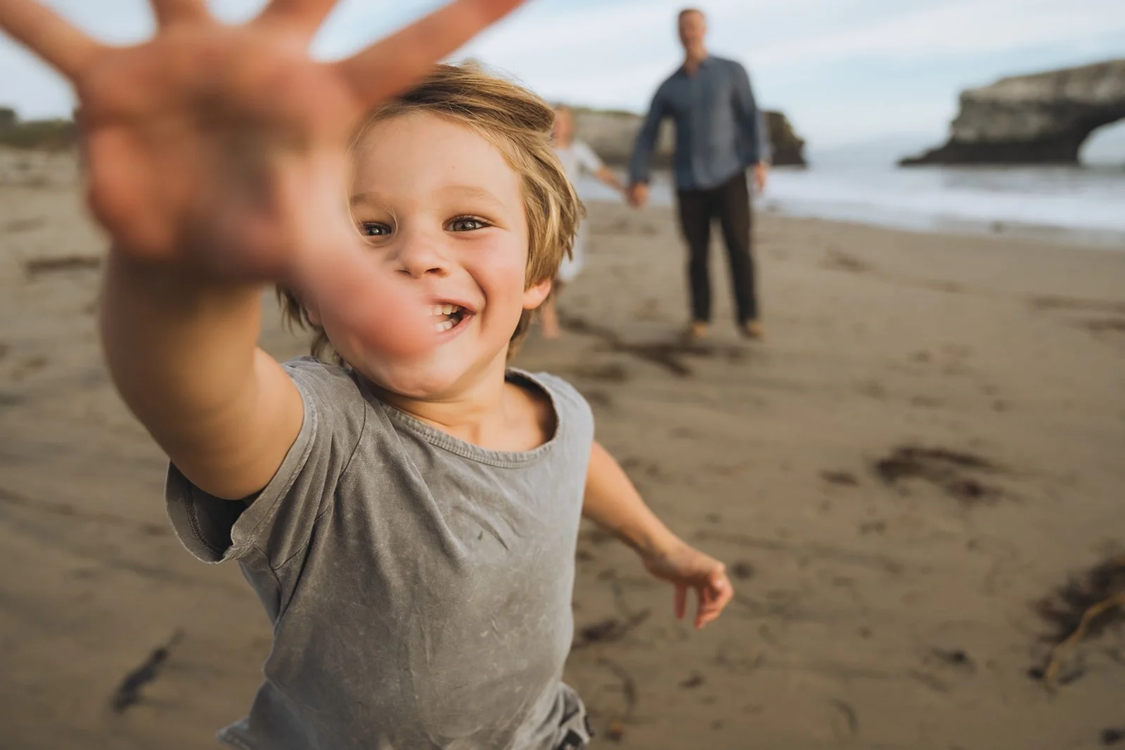 A young boy on a sandy beach reaching out towards the camera, smiling with sandy hands, with two people holding hands and walking along the beach in the background, and rocky formations in the distance.