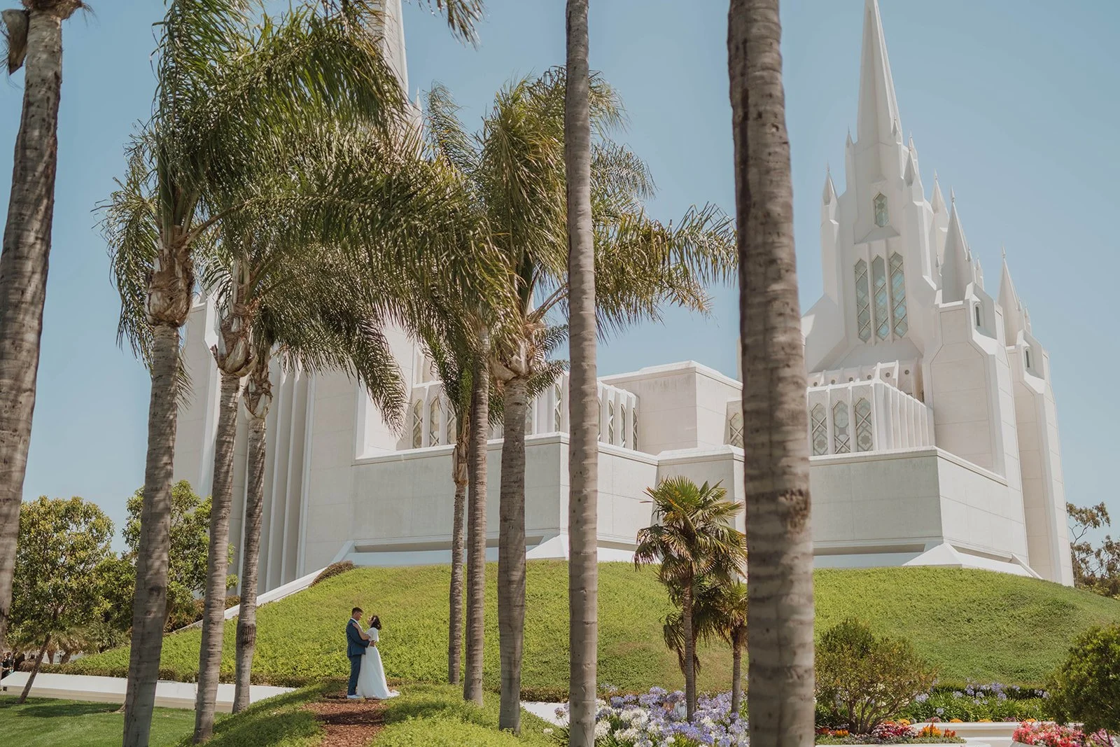 A bride and groom standing and embracing among palm trees on a landscaped lawn, with a large white church or cathedral in the background under a clear blue sky.