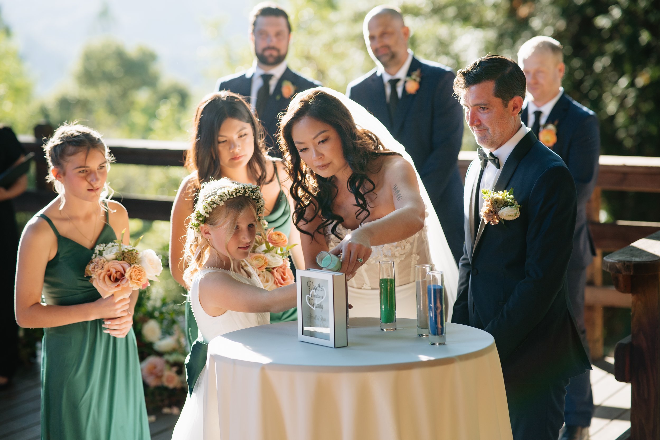 A bride and groom participating in a sand ceremony at their wedding, surrounded by bridesmaids and groomsmen outside on a sunny day.