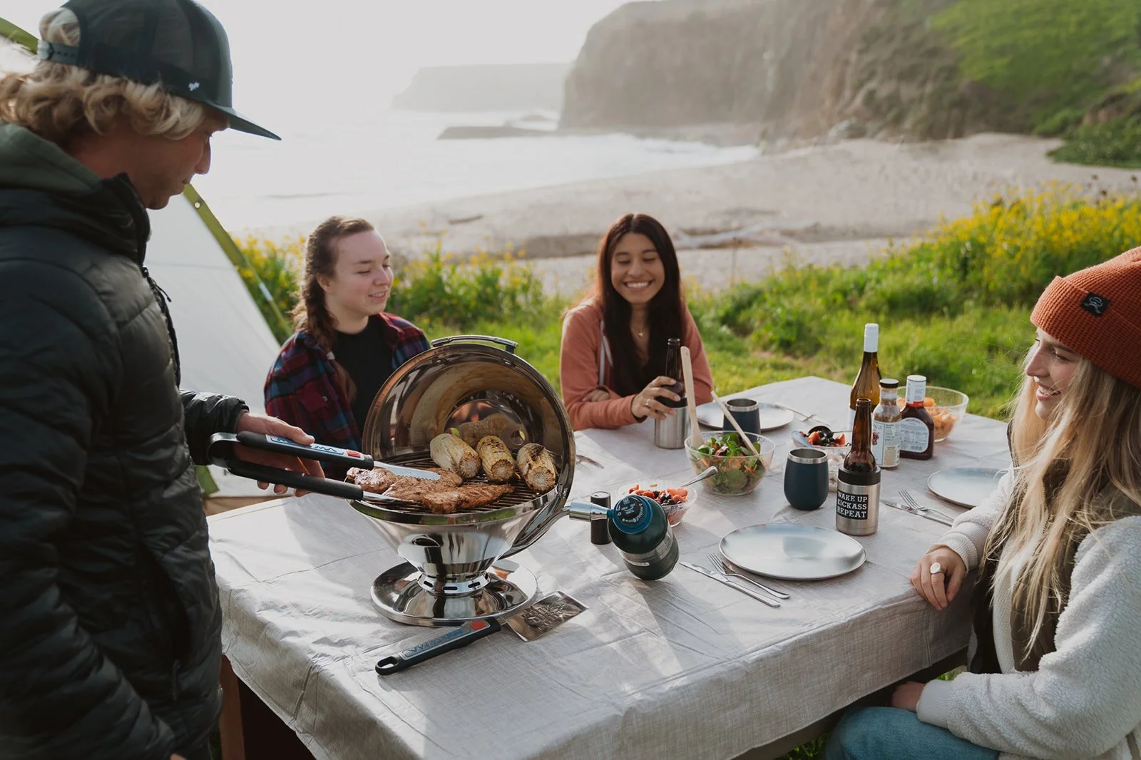 People enjoying a meal outdoors at a table with food and drinks near a beach, with cliffs and greenery in the background.
