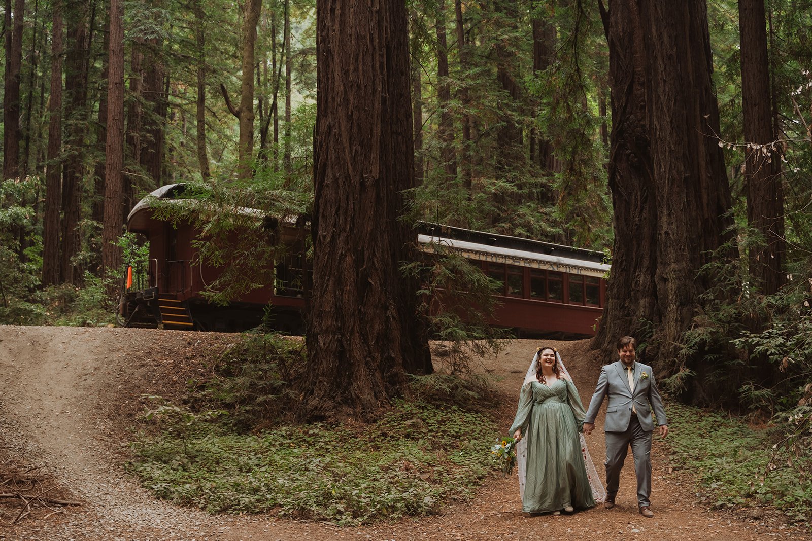 A couple dressed in wedding attire walking hand in hand on a dirt trail in the forest, with tall trees and a red train car in the background.