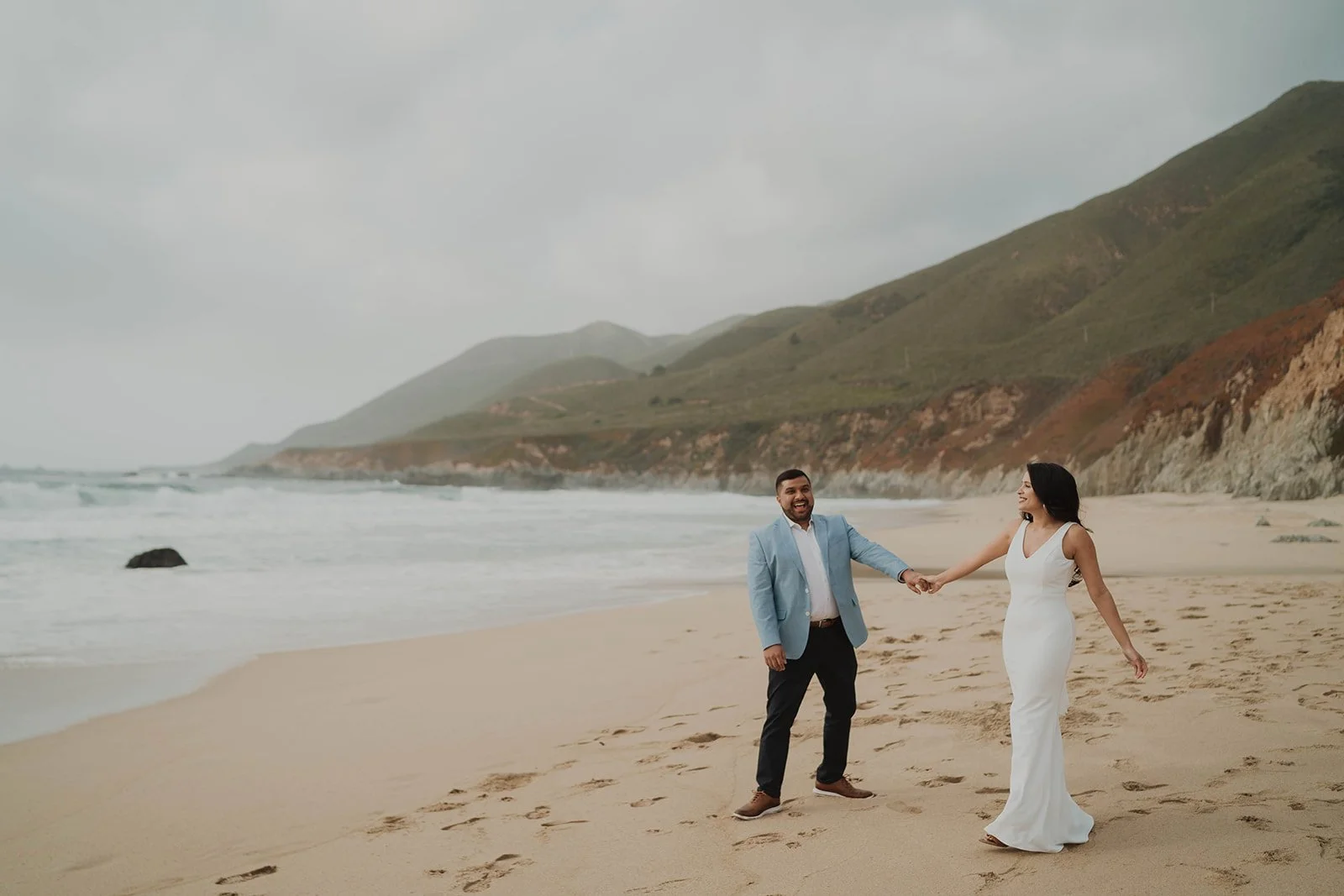 A smiling man and woman holding hands on a beach with ocean waves, rocky shoreline, and green hills in the background.