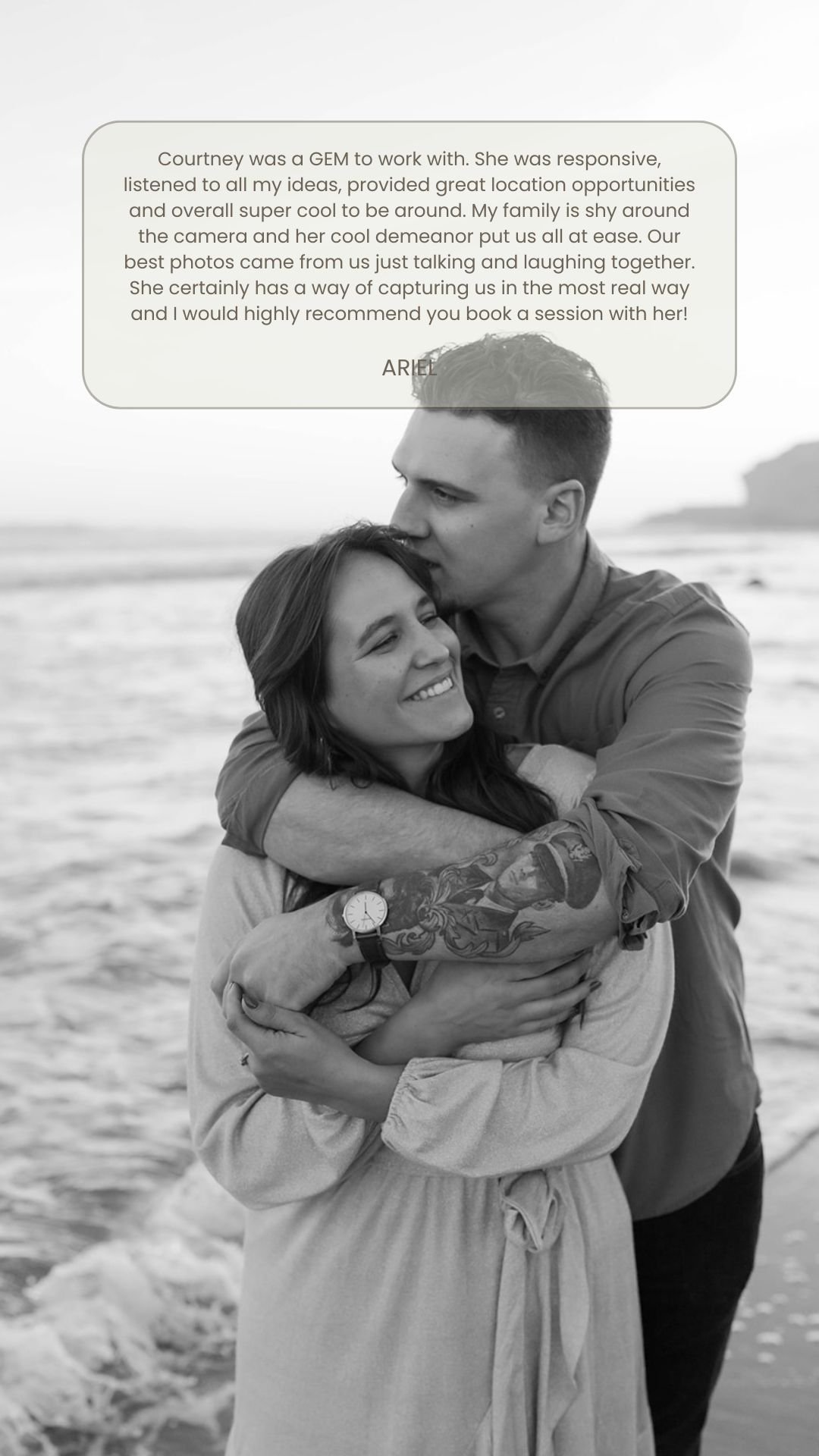 Black and white photo of a couple hugging on the beach, smiling, with water and rocks in the background. The woman has long hair, and the man has short hair and tattoos on his arm.