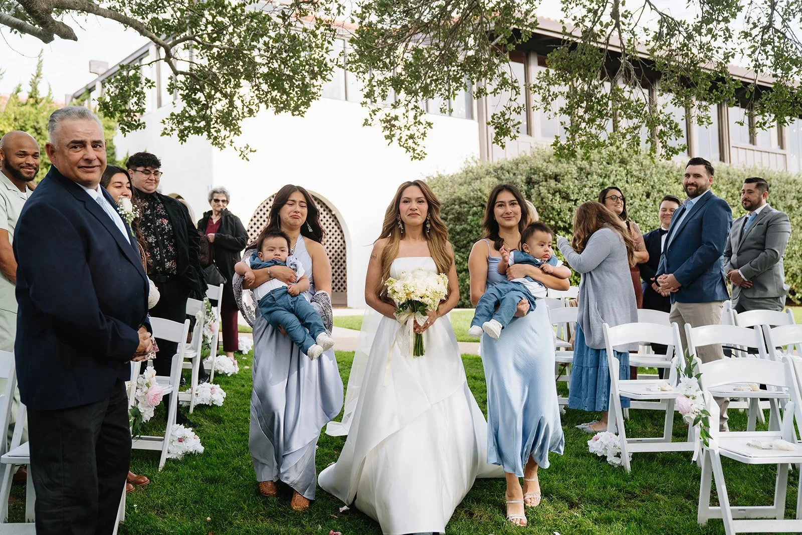Bride walking down an outdoor aisle holding a bouquet, surrounded by family and friends at a wedding ceremony.