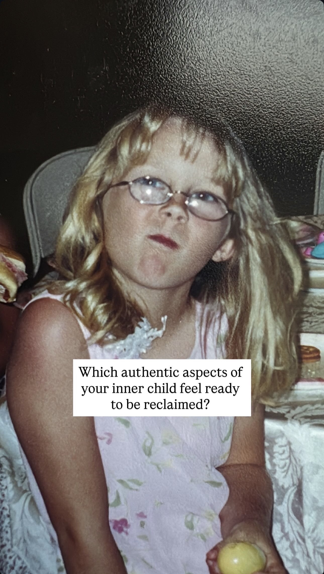 A young girl with blonde hair and glasses sitting at a table, holding a yellow ball, with a serious expression, wearing a white dress with a floral pattern.