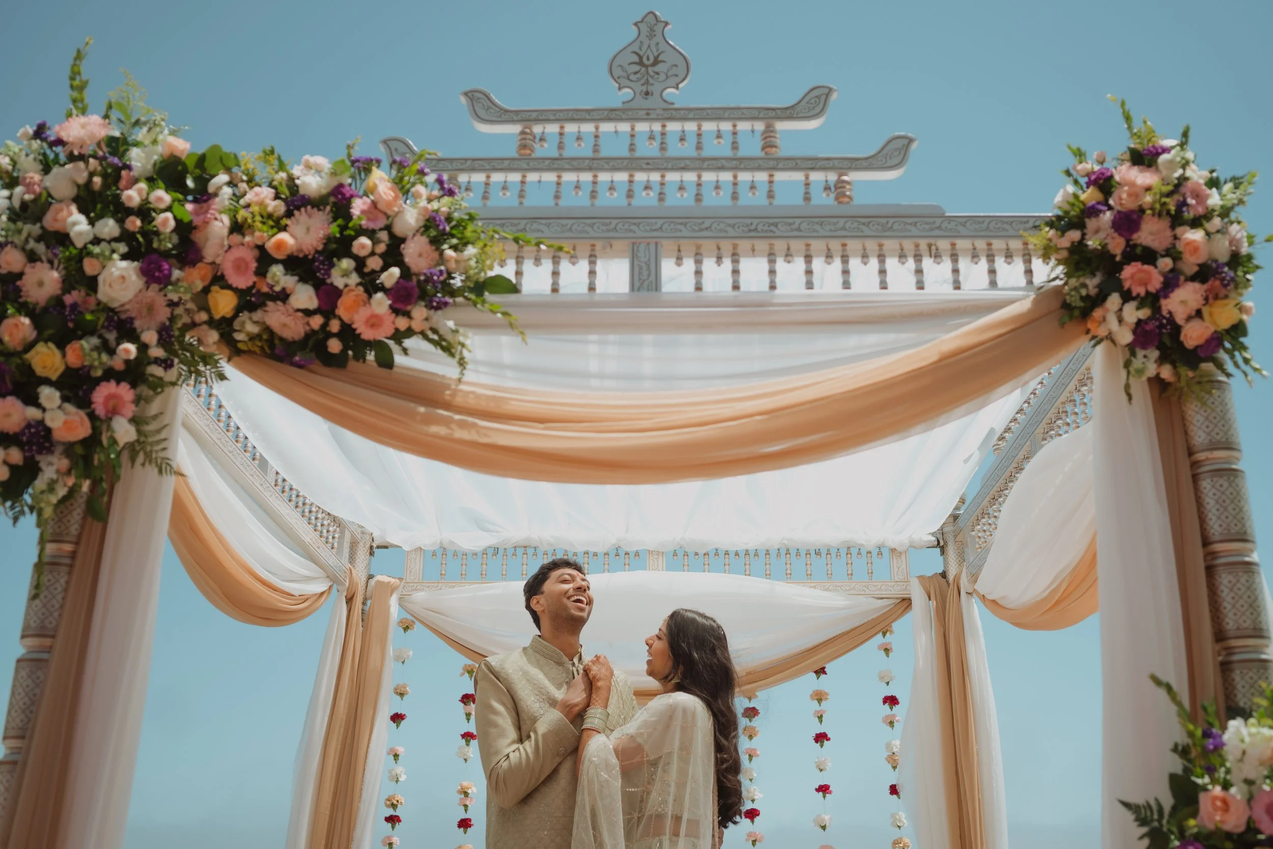 A couple smiling and holding hands under a decorated wedding arch with flowers and fabric outdoors on a sunny day.