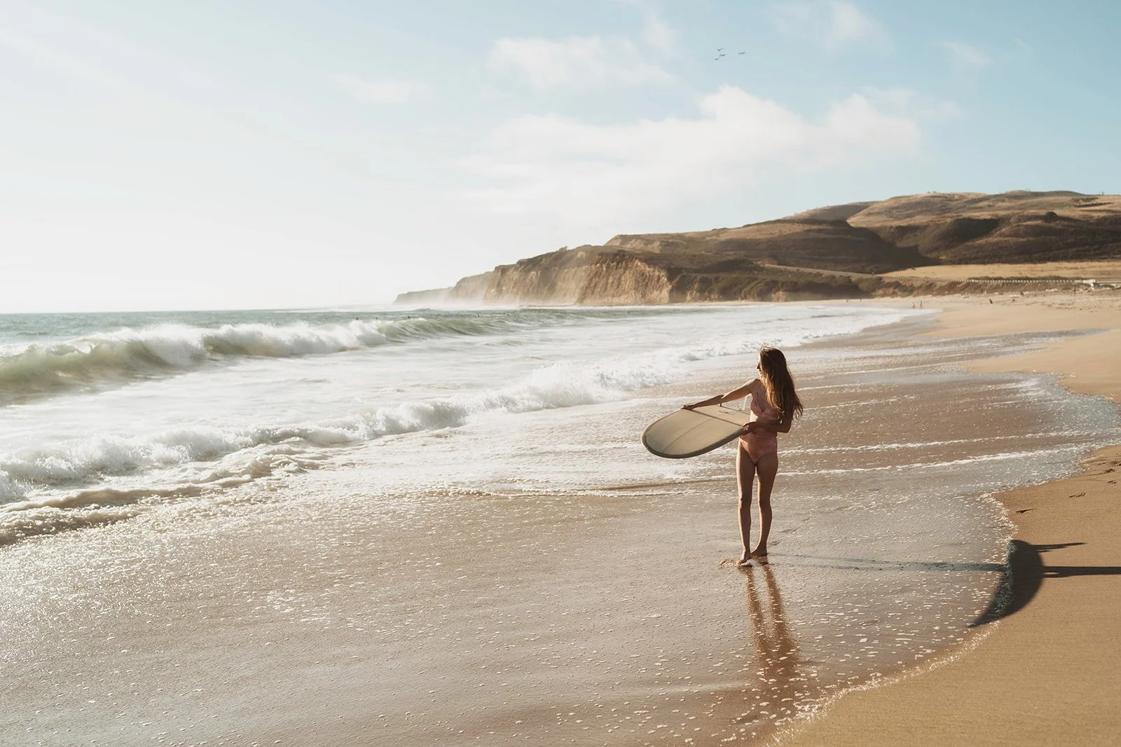 A young woman in a pink swimsuit holding a surfboard stands on the sandy beach near the shoreline, looking towards the ocean with waves and cliffs in the background.