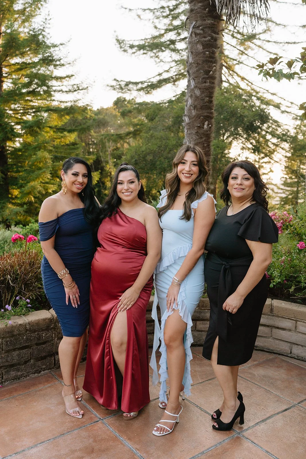 Four women standing outdoors on a patio, dressed in formal dresses. They are smiling and posing together with trees and plants in the background during sunset.