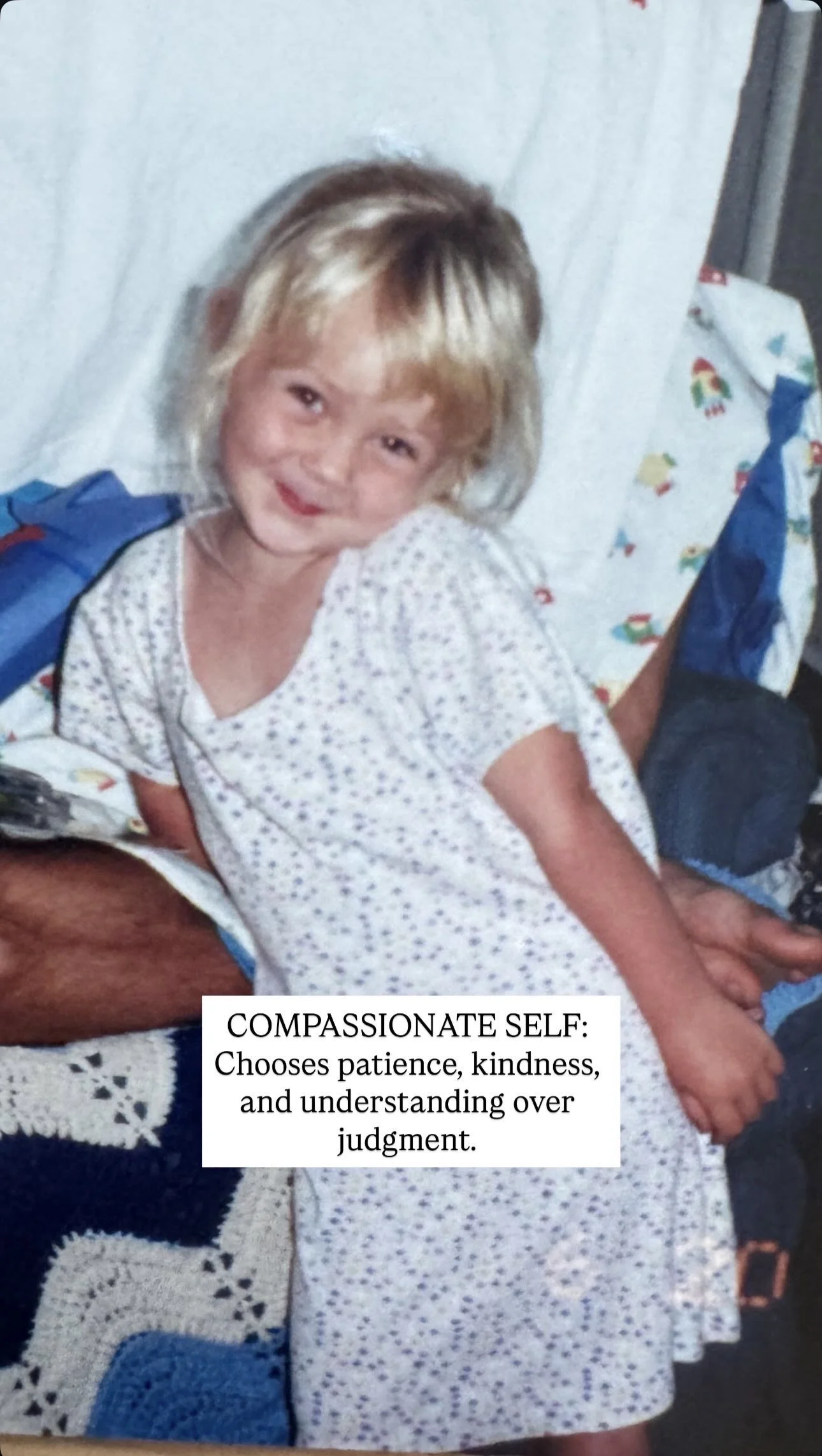 A young girl with blonde hair and a smiling face, wearing a hospital gown with a small floral print, sitting on someone's lap, with a blanket and some bags around her in a room.