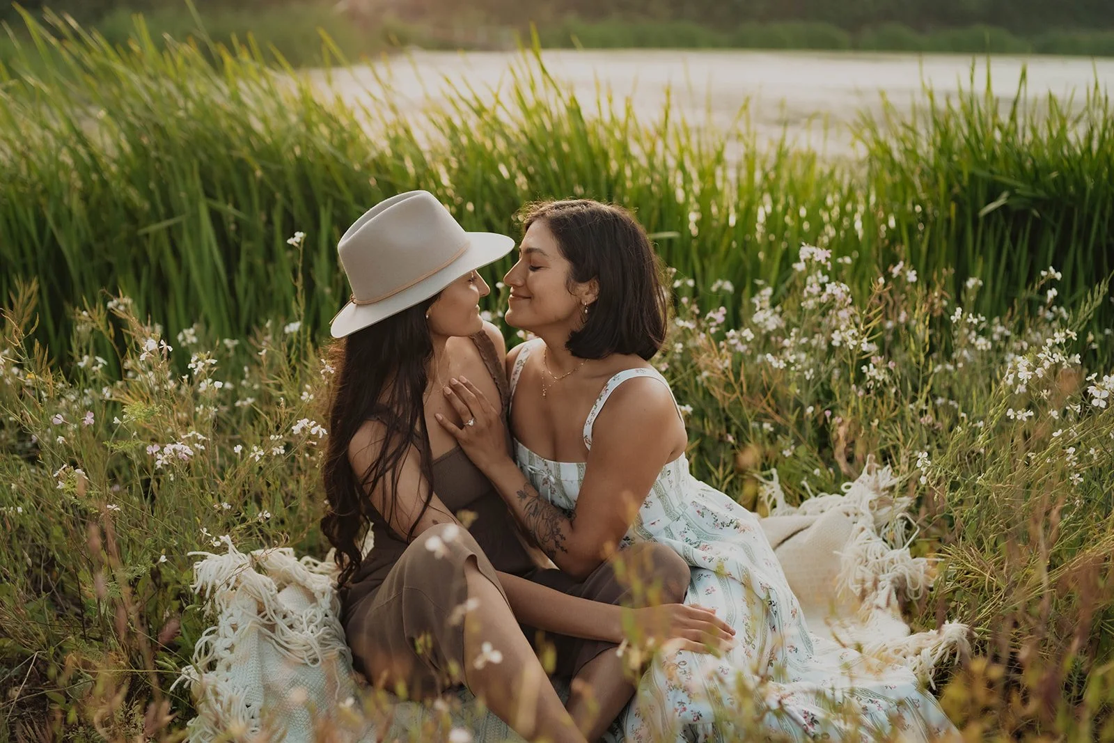 Two women sitting close together in a field of grass and small wildflowers, with a body of water and trees in the background, sharing an intimate moment with their foreheads touching and smiling.